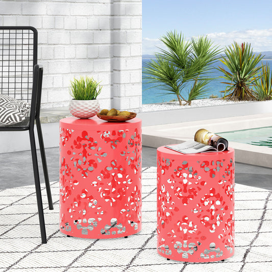 Two Outdoor Metal Modern Design End Tables in coral with cutout patterns sit on a patterned rug by the pool. One table holds a plant and bowl, the other a magazine. Palm trees and the ocean are visible in the background.