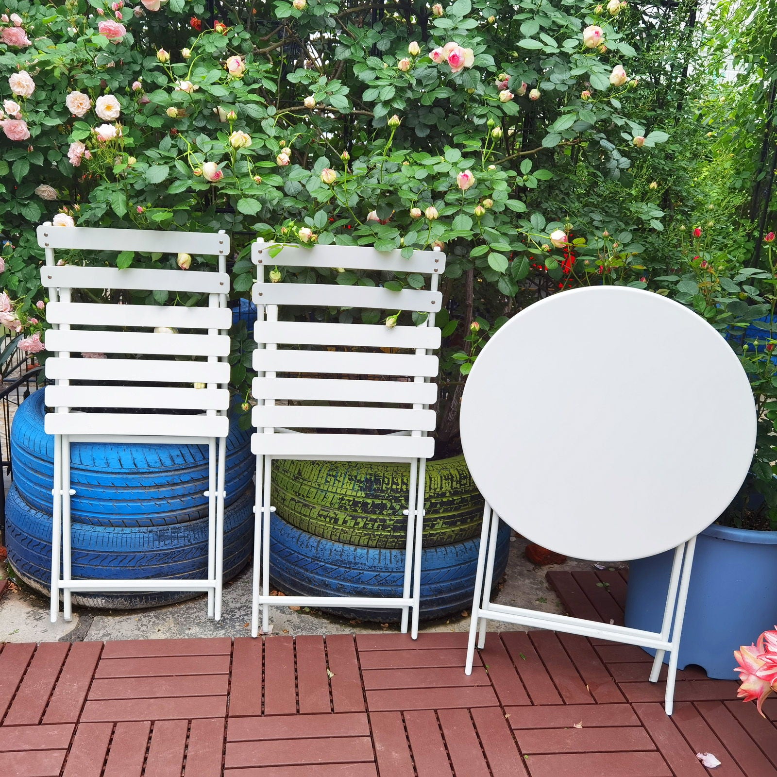 A 3 Piece Patio Bistro Set of foldable round table and chairs is arranged on a red-tiled patio, set against blooming rose bushes and blue and green painted tires repurposed as planters.