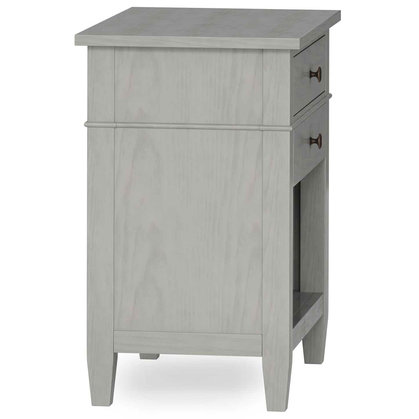 The Carlton Bedside Table, handcrafted in light gray wood, features a single drawer with round metal handles, tapered legs, and an open shelf. Photographed at an angle showing both its side and front.