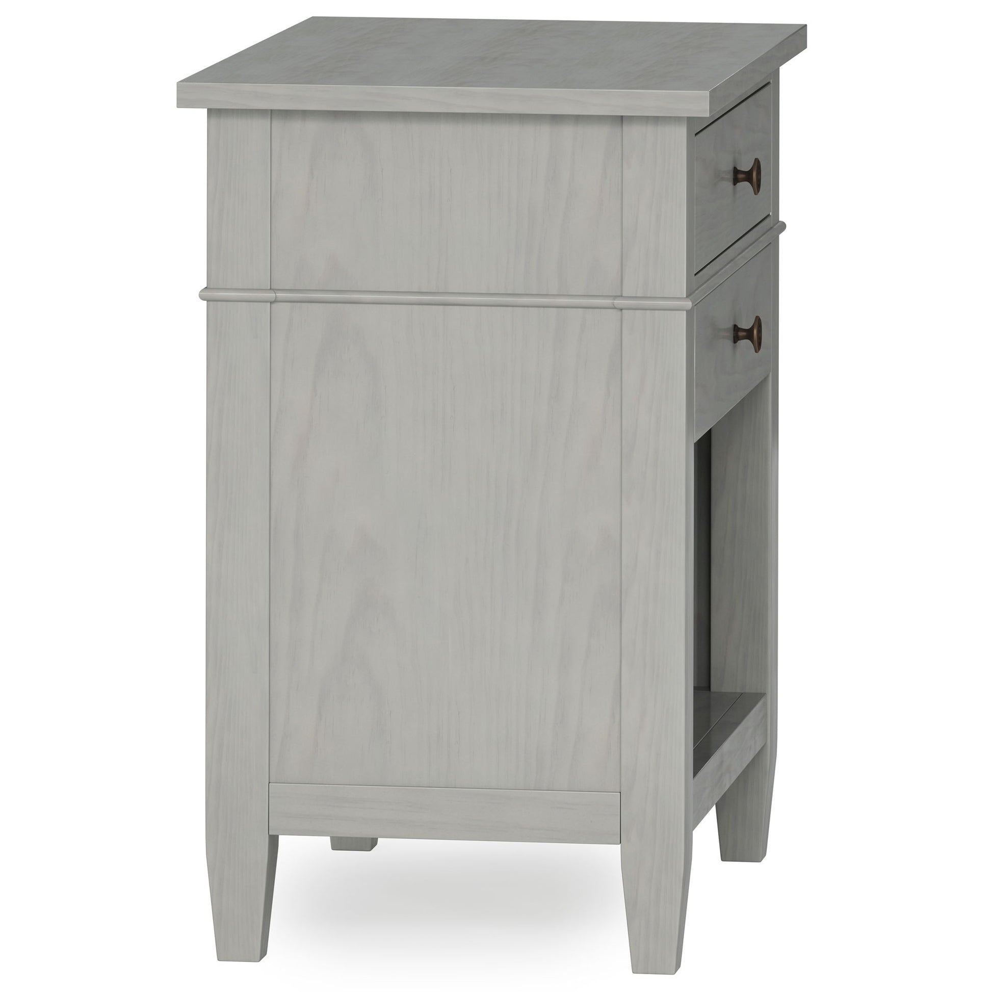 The Carlton Bedside Table, handcrafted in light gray wood, features a single drawer with round metal handles, tapered legs, and an open shelf. Photographed at an angle showing both its side and front.