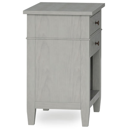 The Carlton Bedside Table, handcrafted in light gray wood, features a single drawer with round metal handles, tapered legs, and an open shelf. Photographed at an angle showing both its side and front.