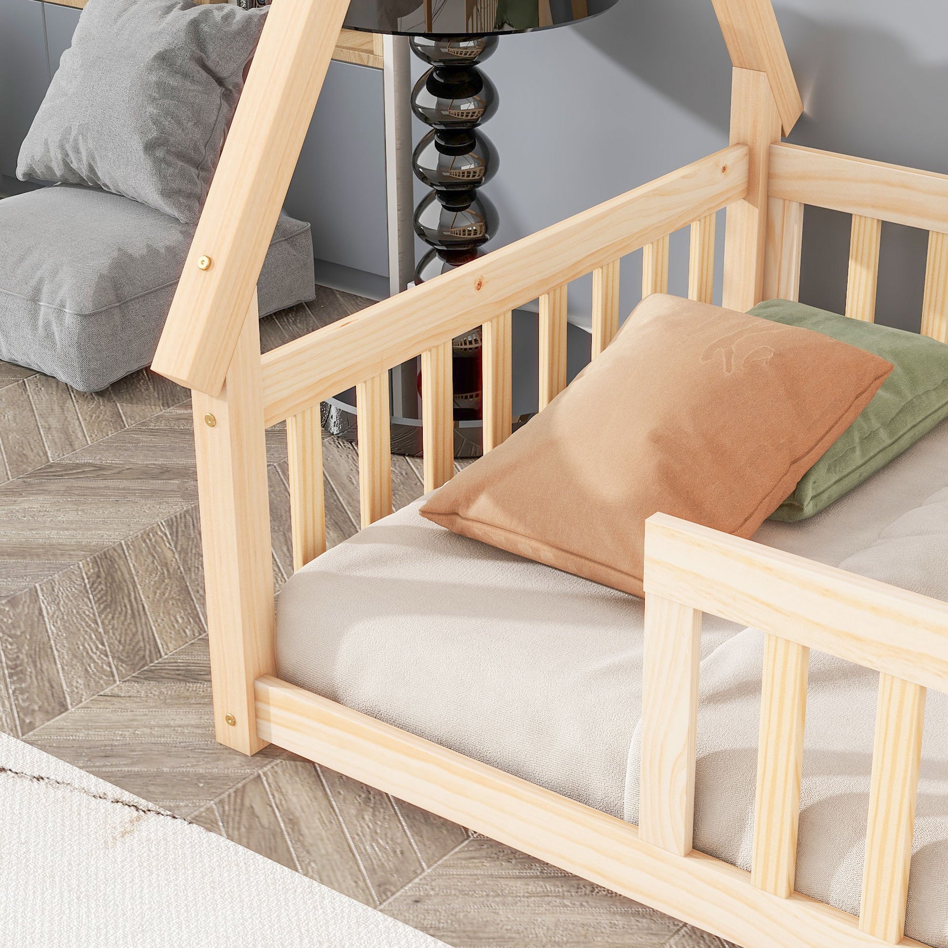 A Tent-Shaped Floor Bed with guardrails and slats, dressed in beige bedding with tan and green pillows, is placed on a patterned wood floor near a black lamp and gray cushion.