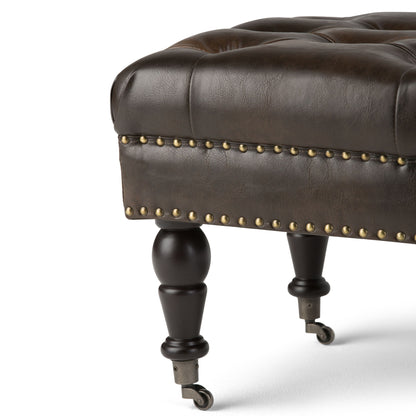 Close-up of the Henley Tufted Ottoman Bench in distressed brown leather, featuring brass nailhead trim, turned wooden legs, and small metal caster wheels, set against a white background.
