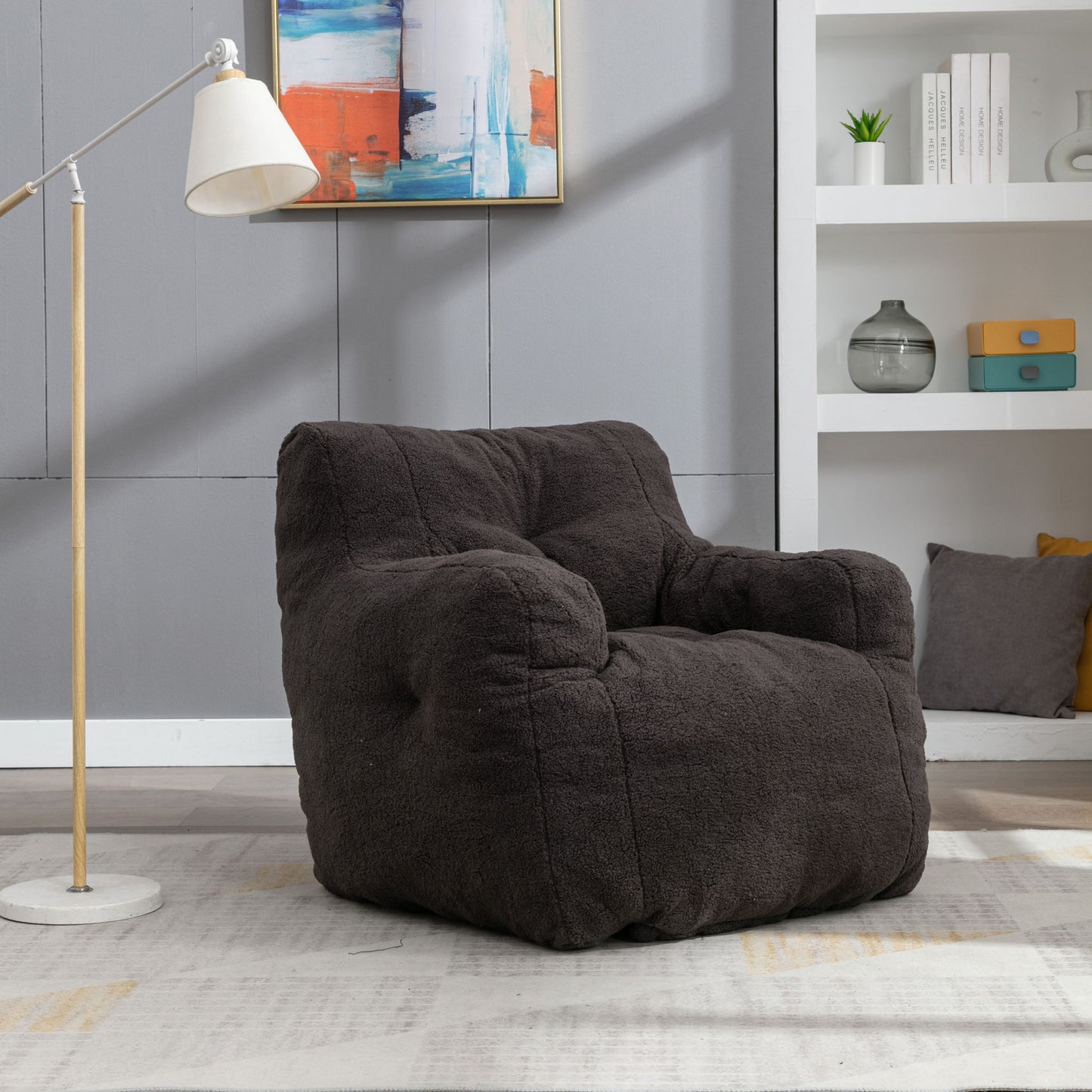 A Soft Teddy Fabric Tufted Foam Bean Bag Chair in black sits on a light-patterned rug in a modern living room, accompanied by a floor lamp, abstract wall art, and shelves filled with books and decorative items.