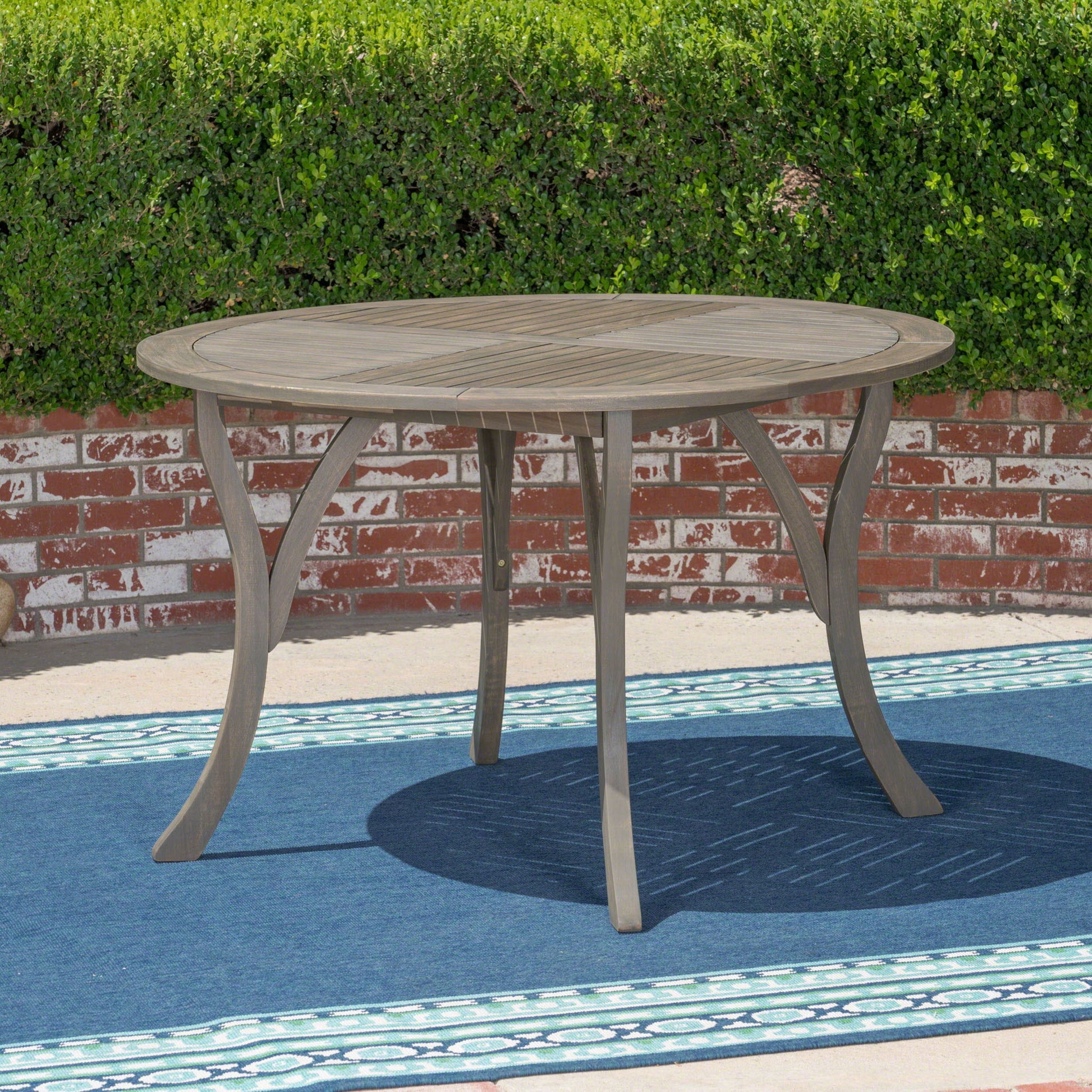 The Hermosa Outdoor Acacia Wood Table, featuring a circular design, stands on a blue patterned rug with a red brick wall and trimmed green hedge in the background.