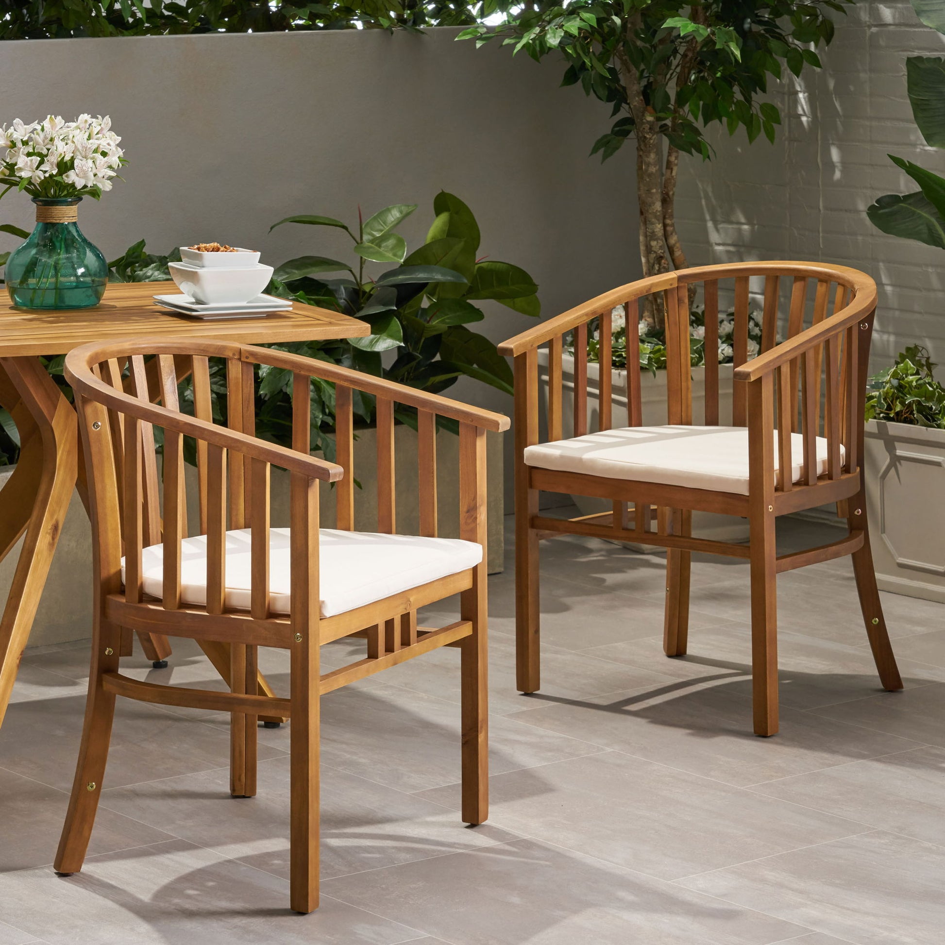 Two Outdoor Wooden Dining Chairs With Cushions (Set of 2) are arranged next to a wooden table with a cup, saucer, and vase of flowers on a tiled patio, surrounded by green plants and a white brick wall.
