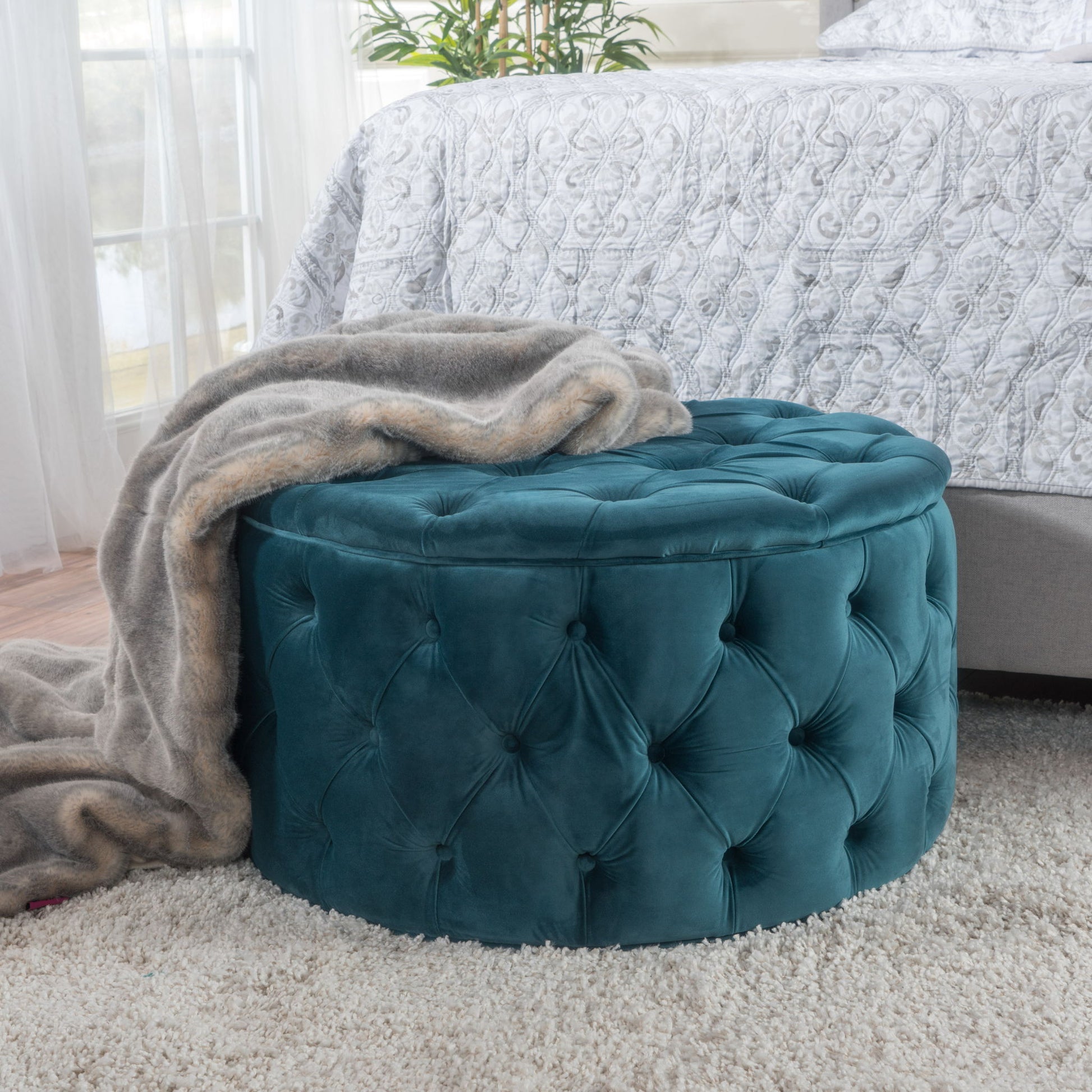 A Modern Glam Ottoman Velvet, round and teal, sits on a cream rug beside a bed, draped with a soft gray faux fur blanket as sunlight streams through the window in the background.
