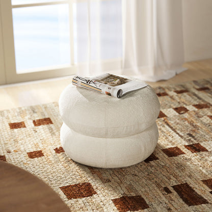 The Nest - Low Round Upholstered Ottoman, featuring a white textured surface, sits on an earth-toned patterned rug by the window. A rolled magazine rests on top as sunlight filters through sheer curtains in the background.