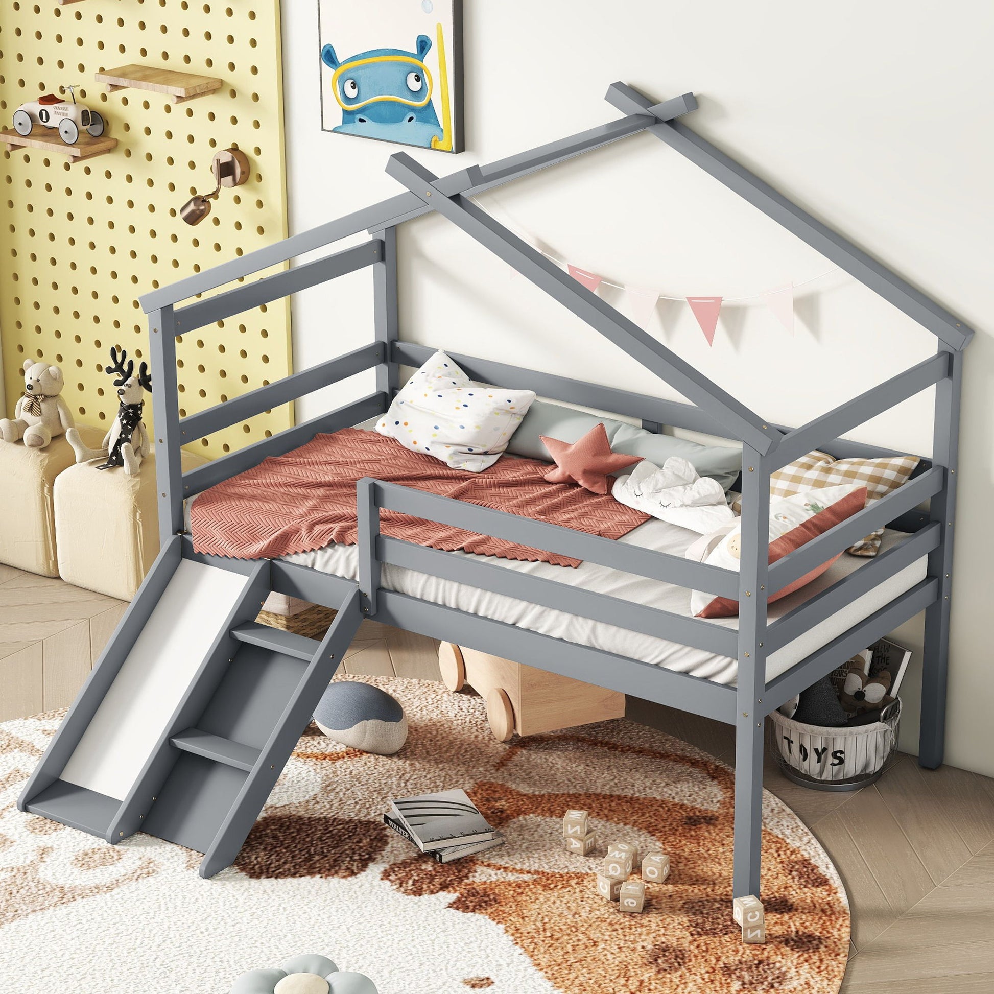 A cozy, playful room features the Low Loft House Bed With Slide, Ladder, Safety Guardrails, and House Roof Frame in gray, styled with soft bedding, pillows, a small banner, stuffed animals, toys on a pegboard, and storage baskets on a beige rug.