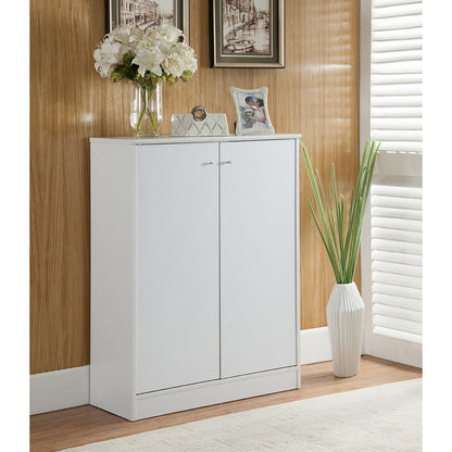 A Shoe / Storage Cabinet With Two Doors Five Shelves stands against a wood-paneled wall, topped with a vase of white flowers, a small clock, and framed photo. Beside it near window blinds is a tall white vase with green leaves.