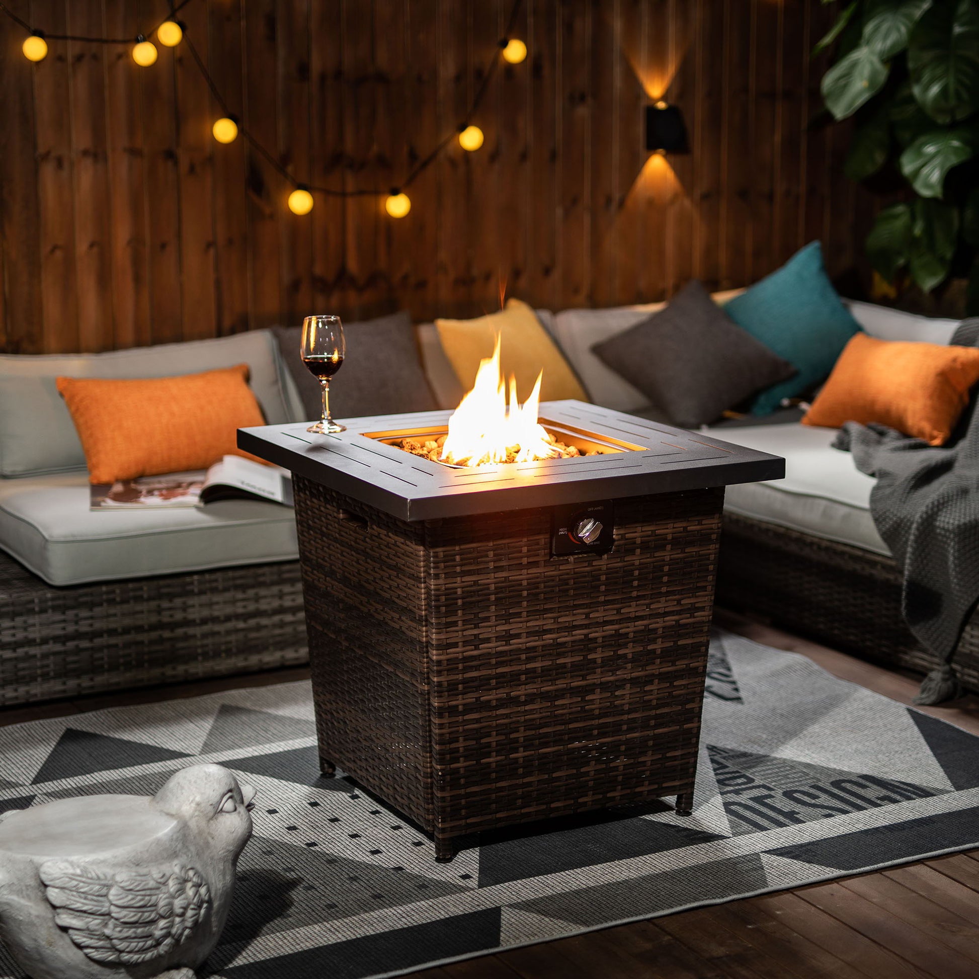 A cozy outdoor patio features the 28" Wicker Square Fire Pit Table - Black Brown lit in the center, surrounded by cushioned seating with orange and gray pillows, string lights overhead, a glass of wine on the table, and a decorative bird statue nearby.