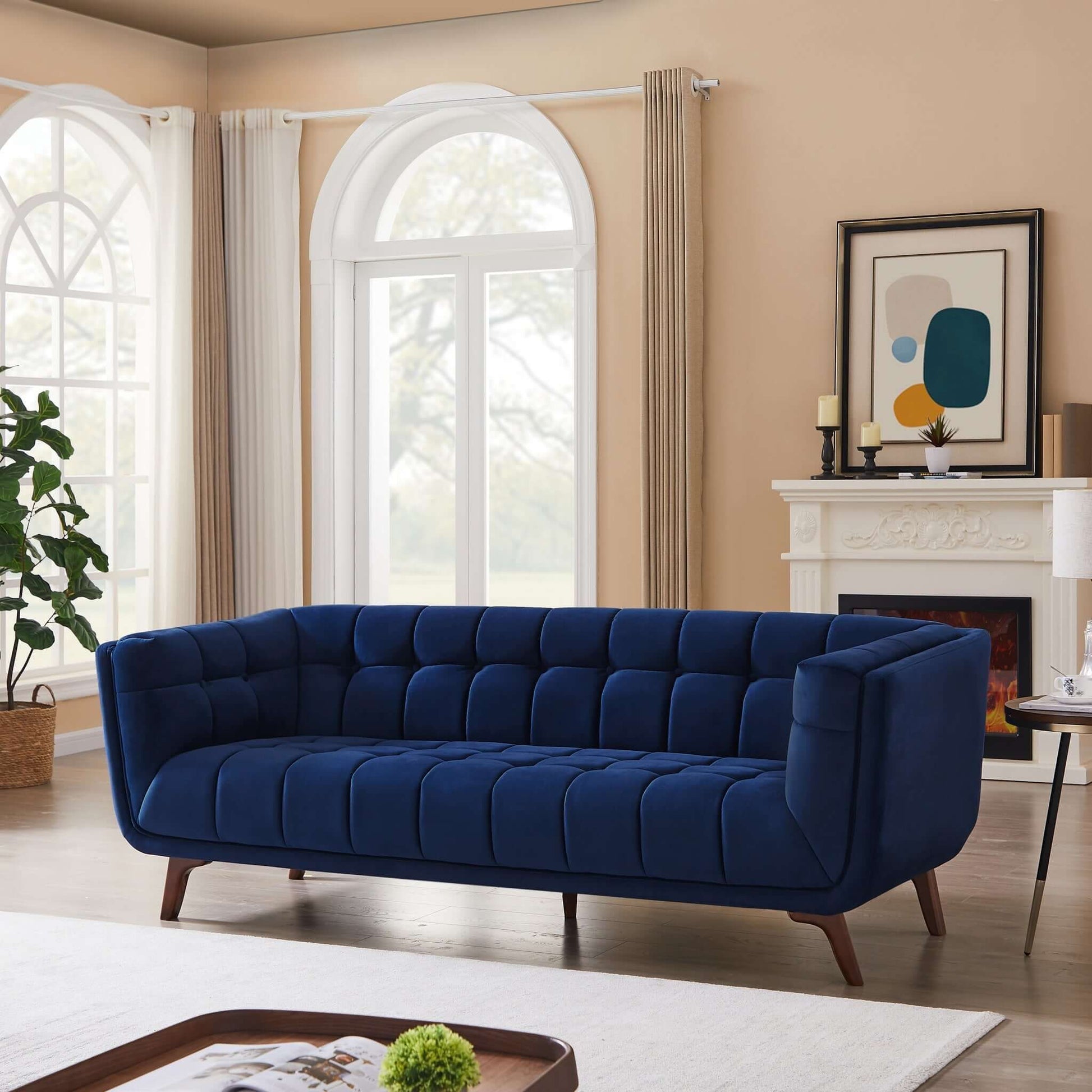 A modern living room with the Addison Mid-Century Modern Tufted Sofa in deep blue, light walls, large arched windows with curtains, a fireplace topped with candles and decor, framed abstract art, a plant, and a beige rug on wood floors.