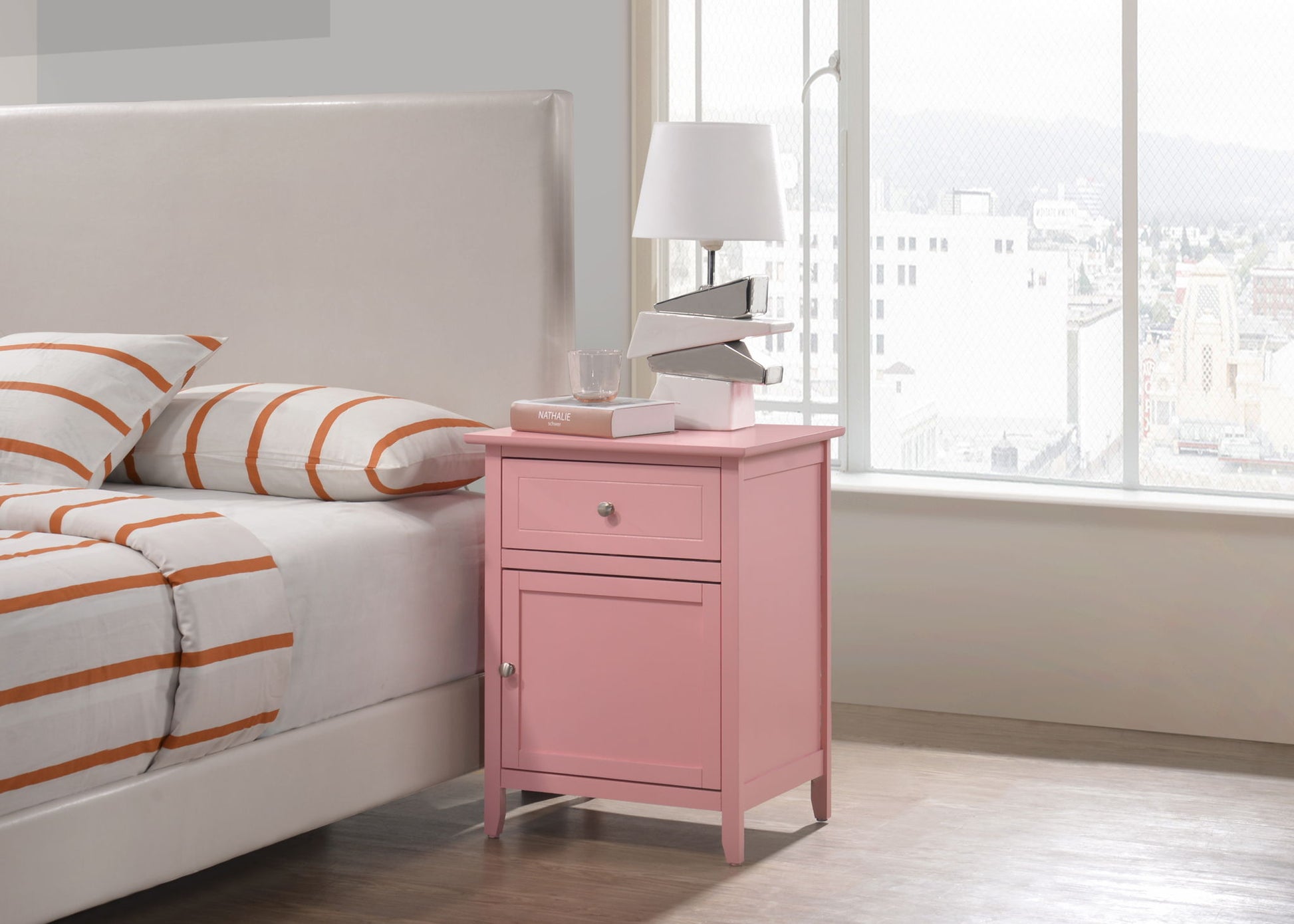 The Izzy - 1 Drawer / 1 Door Nightstand in pink stands by a bed with white and orange striped bedding. On it are a lamp, glass of water, and two books. A large window nearby offers a city view.