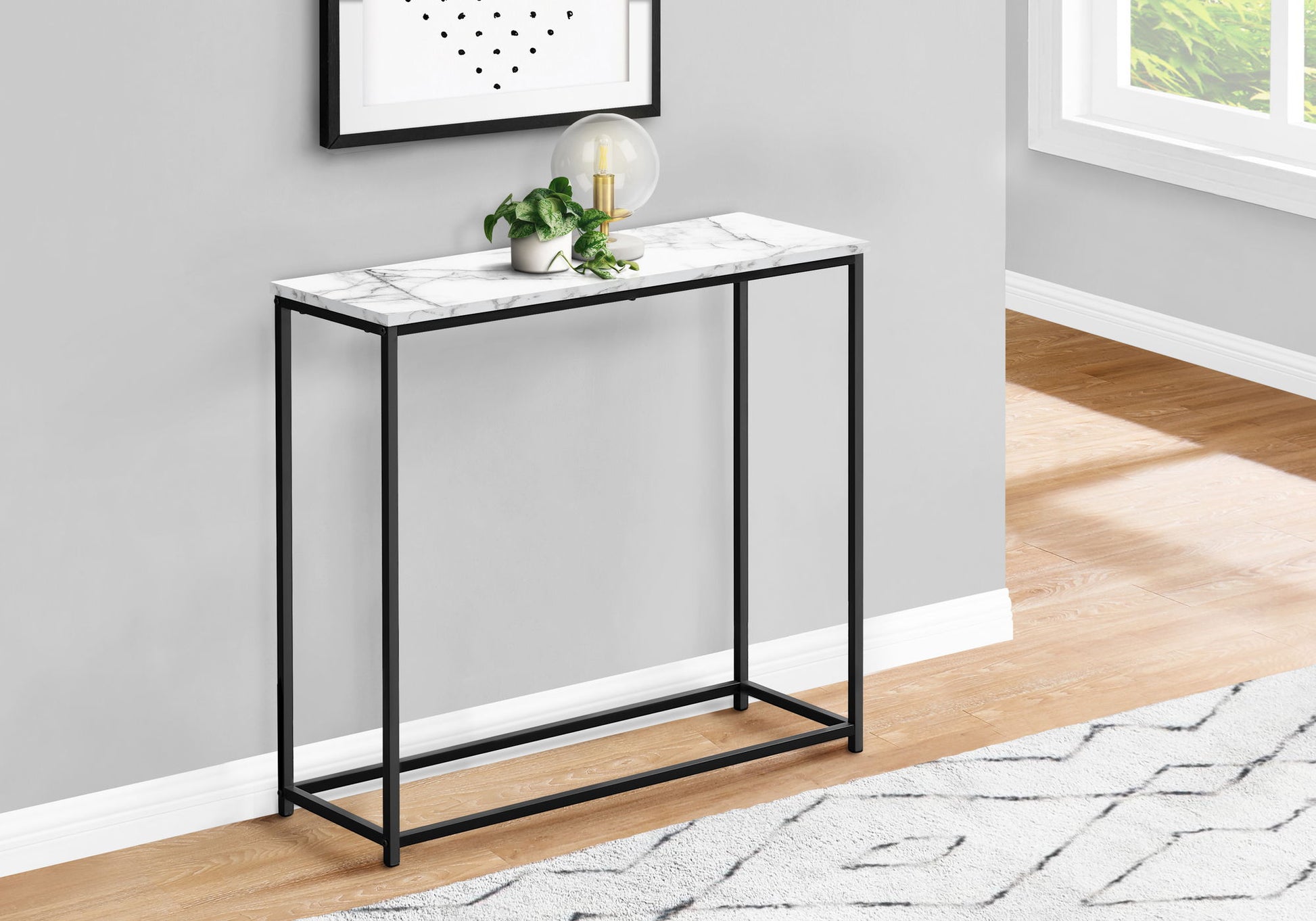 The Accent Console Table For Entryway, featuring a white marble top and black metal frame, stands against a gray wall. A small plant and glass lamp rest on it, with a framed print above and sunlight streaming through a nearby window.