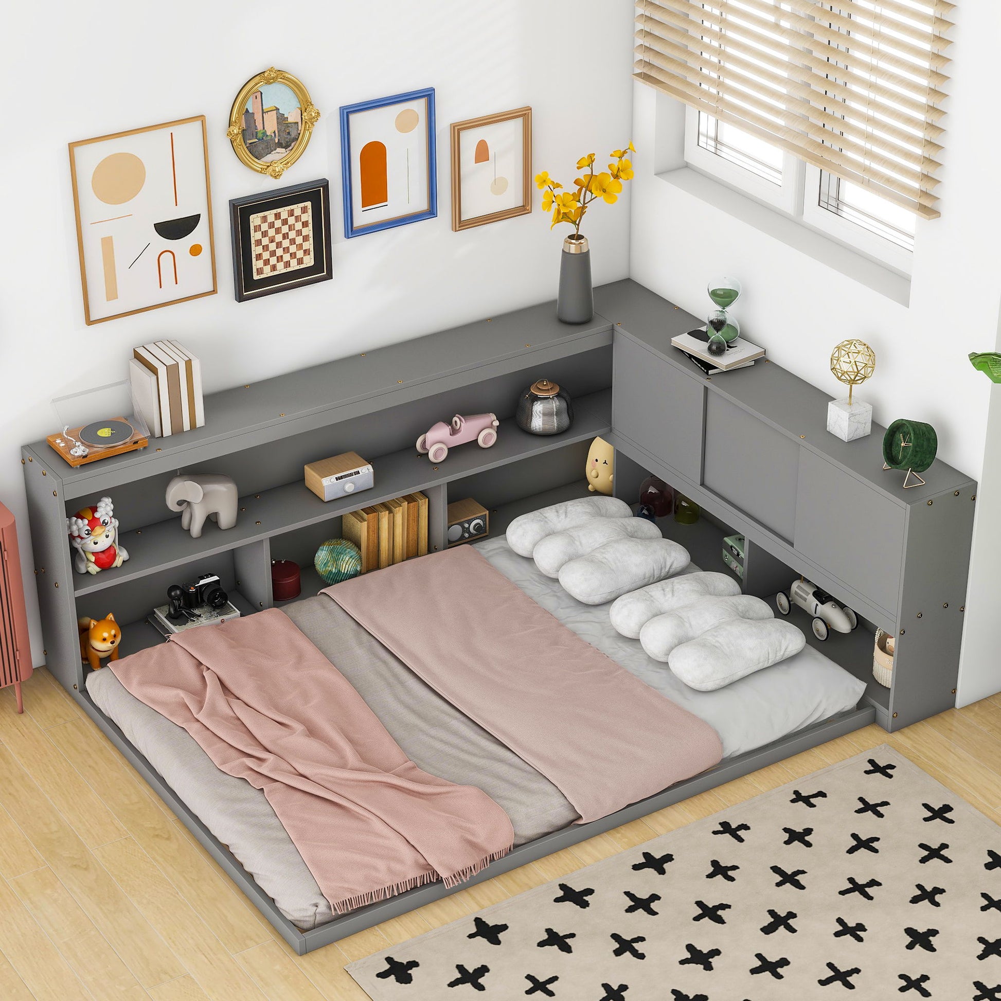 A cozy bedroom corner features the Floor Bed With L-Shaped Bookcases, Sliding Doors, Without Slats, styled with pink and beige bedding, cloud-shaped pillows, toys and decor on the shelves, framed wall art, and a daylight-filled window with blinds.