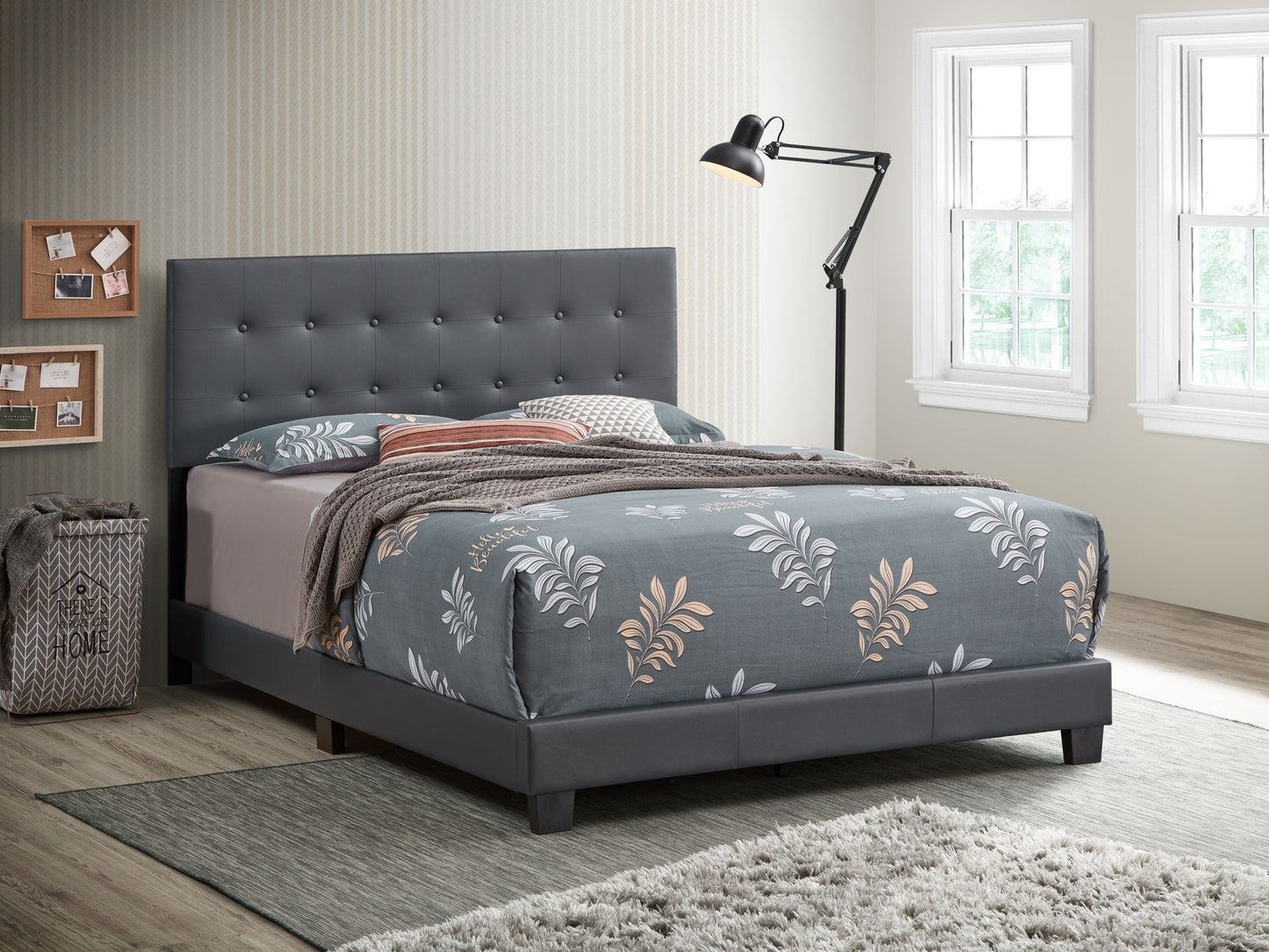 A modern bedroom with the gray Caldwell bed, floral bedding, a black floor lamp, cork boards on the wall, a gray laundry basket, and large windows filling the space with natural light.