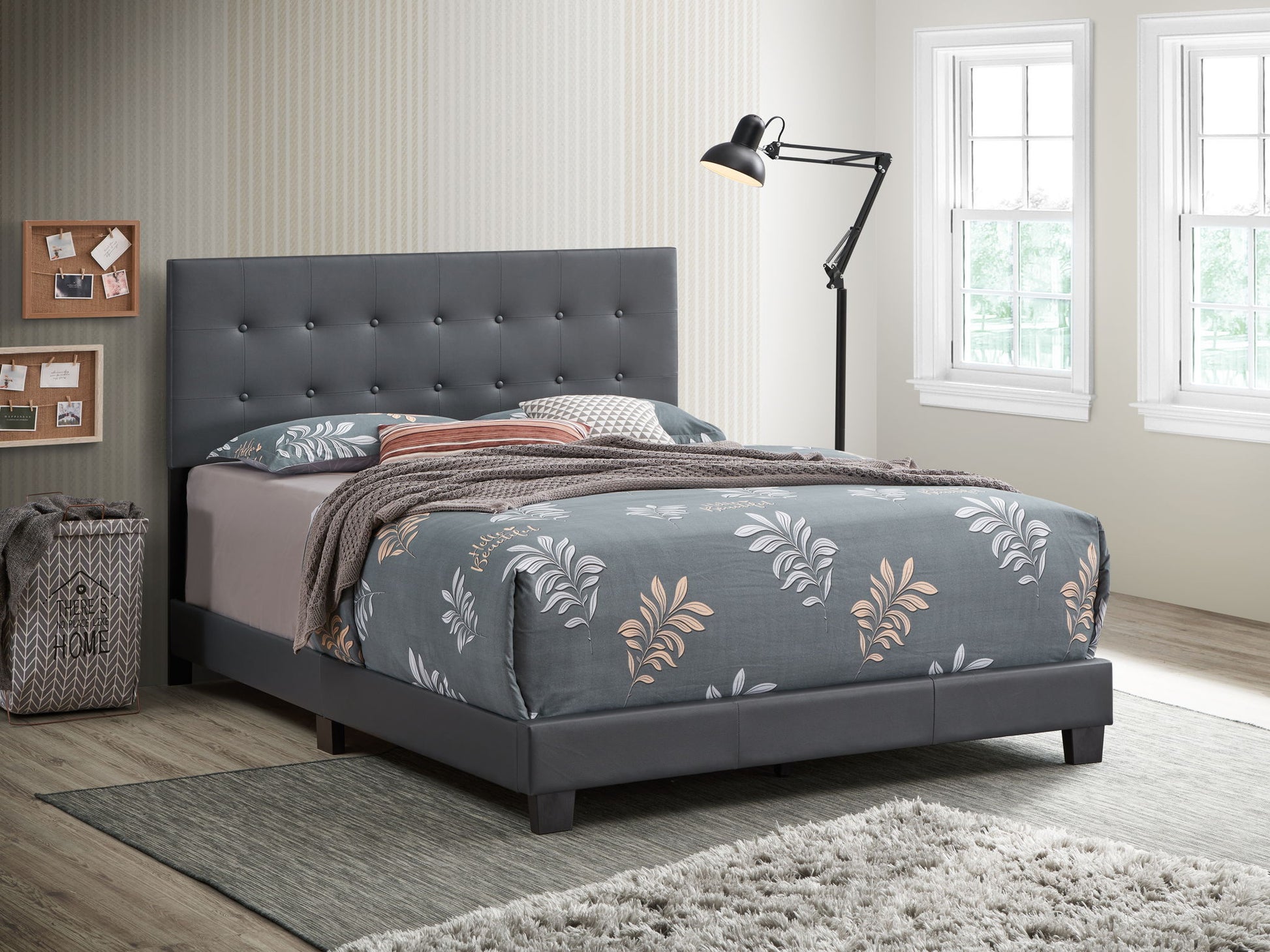 A modern bedroom with the gray Caldwell bed, floral bedding, a black floor lamp, cork boards on the wall, a gray laundry basket, and large windows filling the space with natural light.