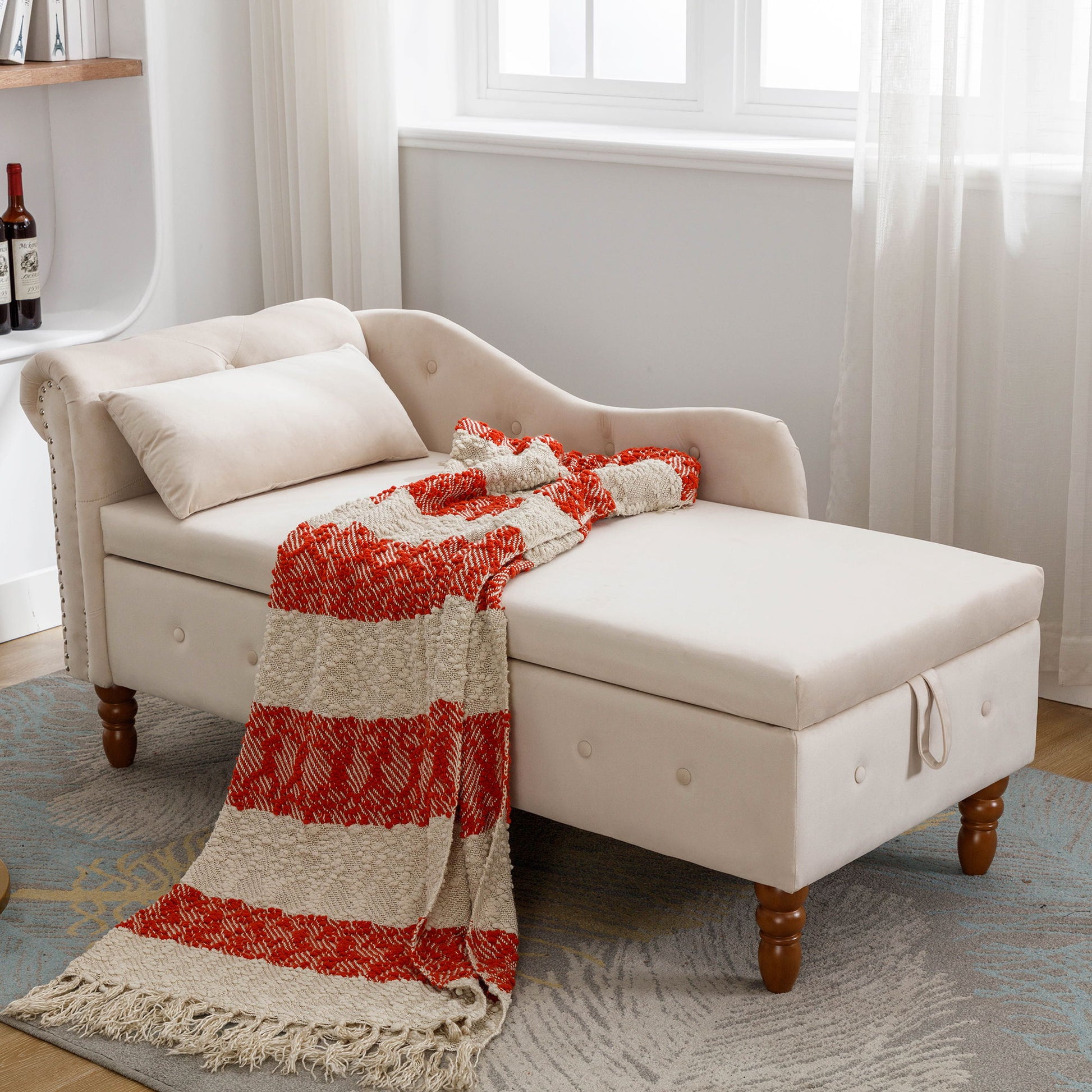 A cream velvet Chaise Lounge Indoor with storage, nailhead trim, rolled arms, and pillow sits by a window. A red and beige striped throw is draped on the seat. Bottles are visible on a shelf in the background.