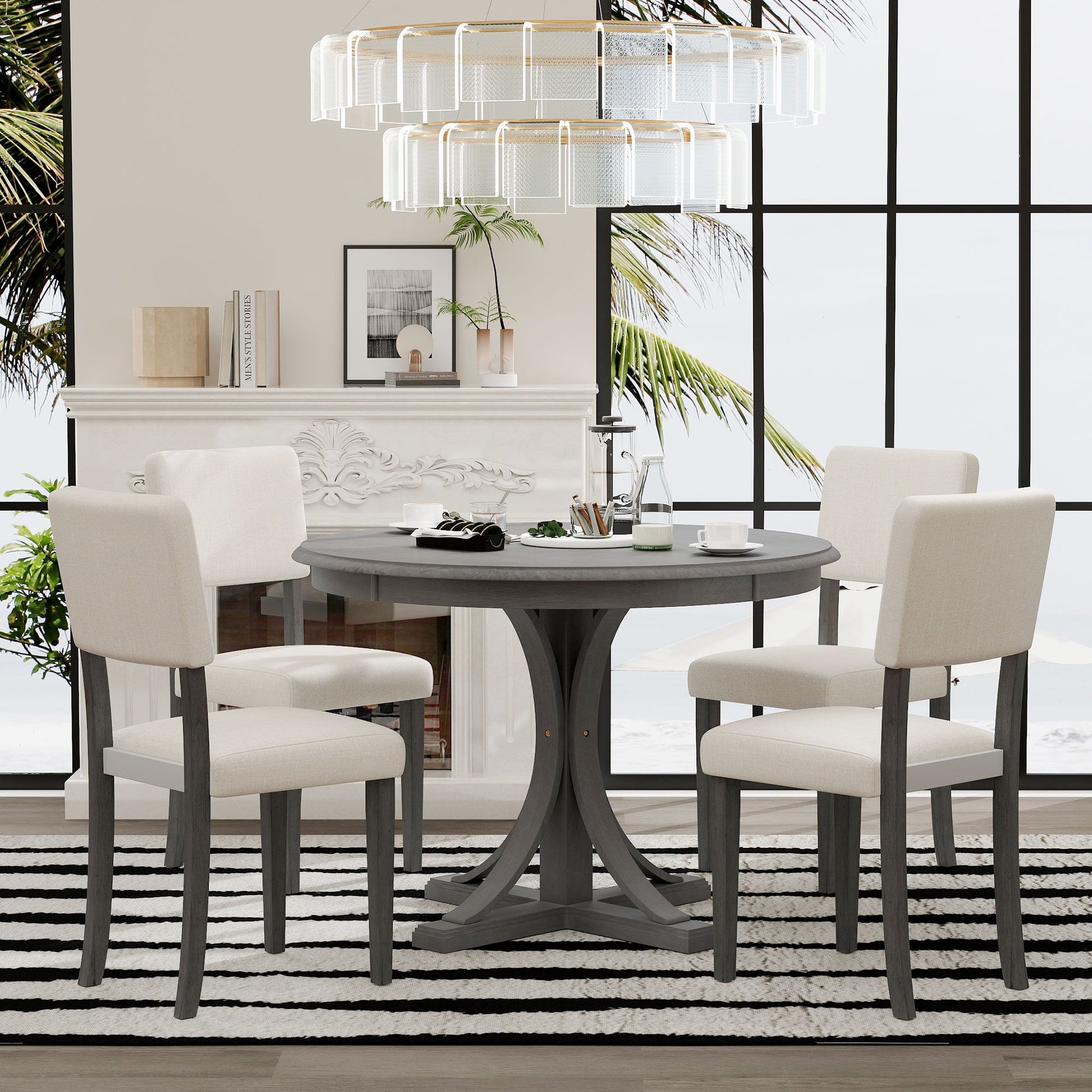 A modern dining room features the Retro Round Dining Table Set with curved trestle legs and four cream upholstered chairs, a striped rug, large chandelier, tall windows with palm views, and neutral decor on the mantel.