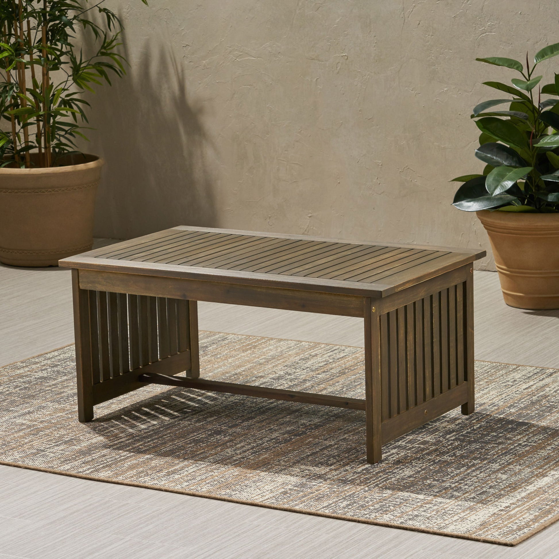 The Acacia Wood Rectangular Patio Coffee Table For Outside sits on a woven rug, surrounded by potted green plants with a beige textured wall in the background.