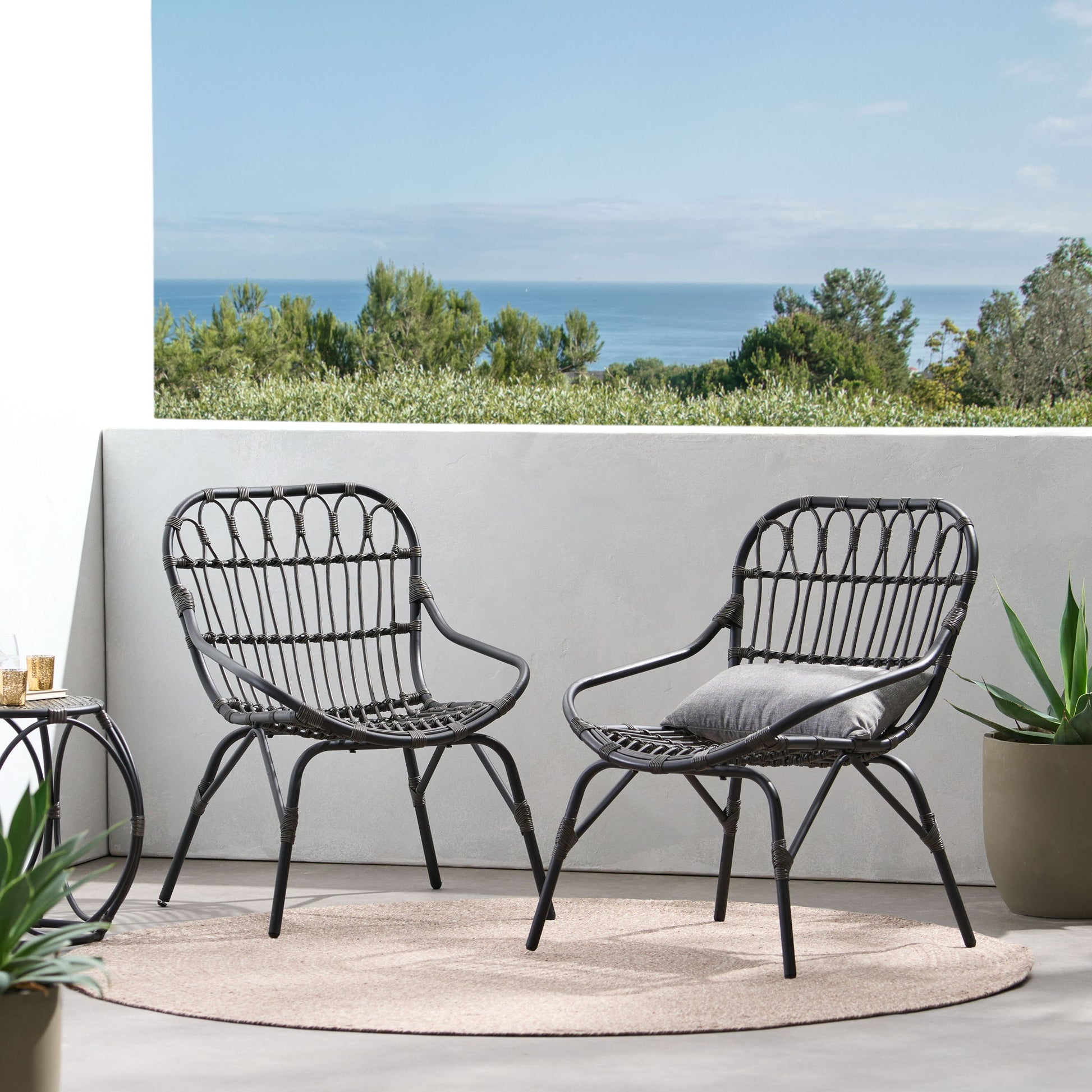 Two Monsoon Outdoor Accent Chairs with open weave design and gray cushions sit on a round beige rug on the patio, surrounded by potted plants with a scenic view of trees and the ocean in the background.