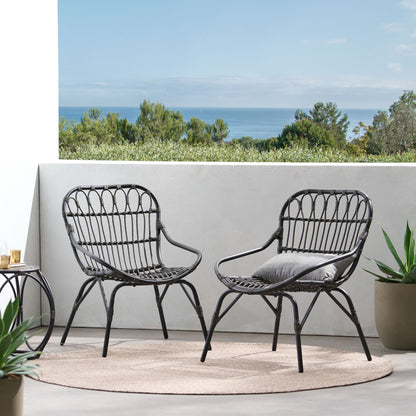 Two Monsoon Outdoor Accent Chairs with open weave design and gray cushions sit on a round beige rug on the patio, surrounded by potted plants with a scenic view of trees and the ocean in the background.