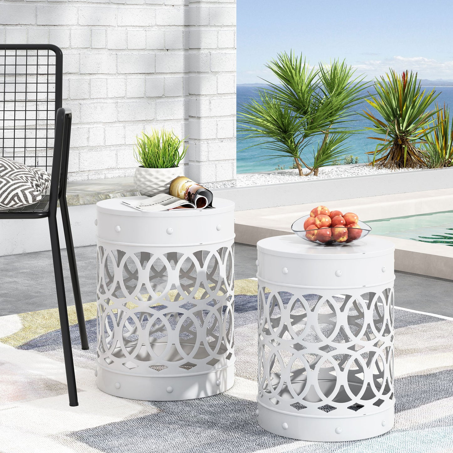 Two Outdoor Metal S/2 End Tables with white finish and circular patterns are arranged poolside overlooking the ocean and palm trees. One holds a bowl of peaches; the other displays a plant, cup, and magazines. A chair is partially visible nearby.
