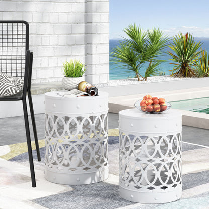 Two Outdoor Metal S/2 End Tables with white finish and circular patterns are arranged poolside overlooking the ocean and palm trees. One holds a bowl of peaches; the other displays a plant, cup, and magazines. A chair is partially visible nearby.