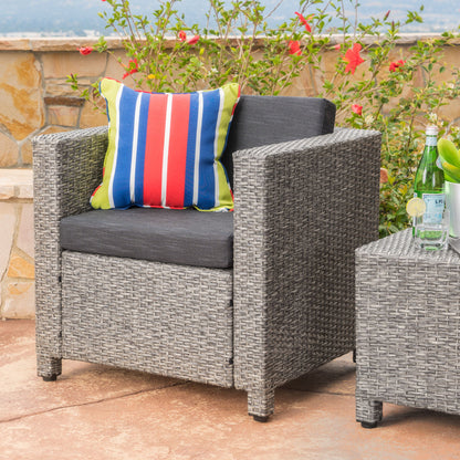 A Stylish Outdoor Club Chair With Weather Resistant Cushions sits on a stone patio with a colorful pillow, next to a matching table holding sparkling water bottles and surrounded by blooming flowers.