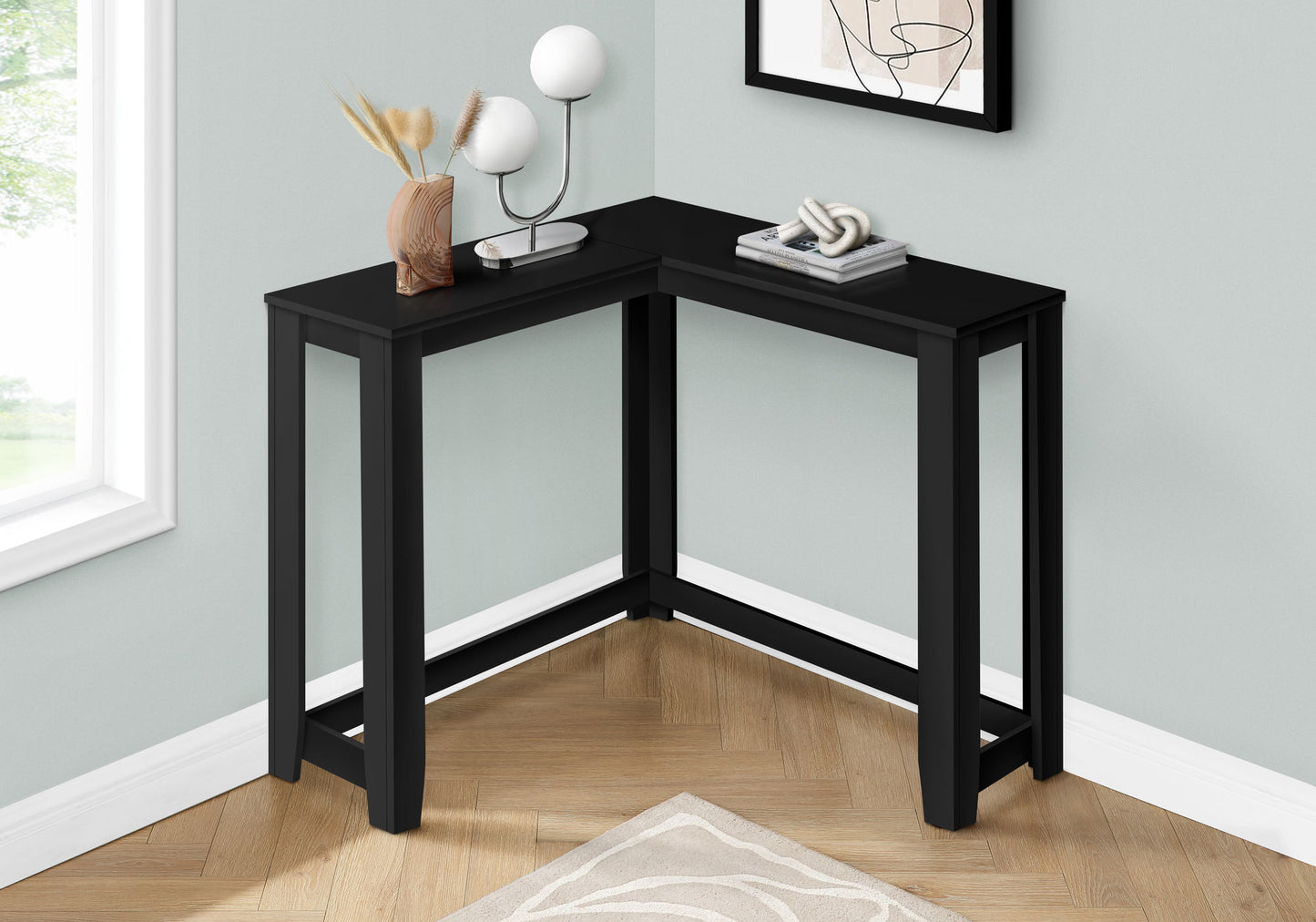A Corner Accent Console Table For Entryway, featuring a unique contemporary design in black, is styled with a lamp, dried plants in a vase, a book, and an abstract sculpture. It sits against pale gray walls on a wooden floor with a light rug.