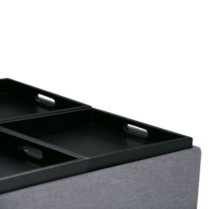 Close-up of the Avalon Square Coffee Table Storage Ottoman, featuring black rectangular trays with handles, placed on a light gray upholstered surface against a white background.
