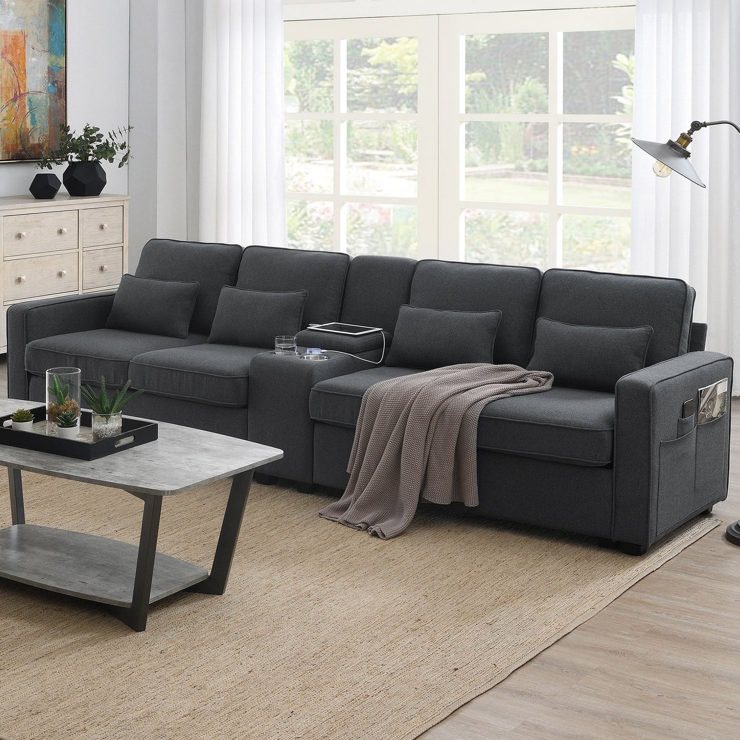 A modern living room with a dark gray upholstered 4-seat sofa featuring a console, 2 cupholders, 2 USB ports (wired/wireless charging), modern linen fabric, 4 pillows, a beige throw blanket, gray coffee table, floor lamp, dresser, and garden-view windows.