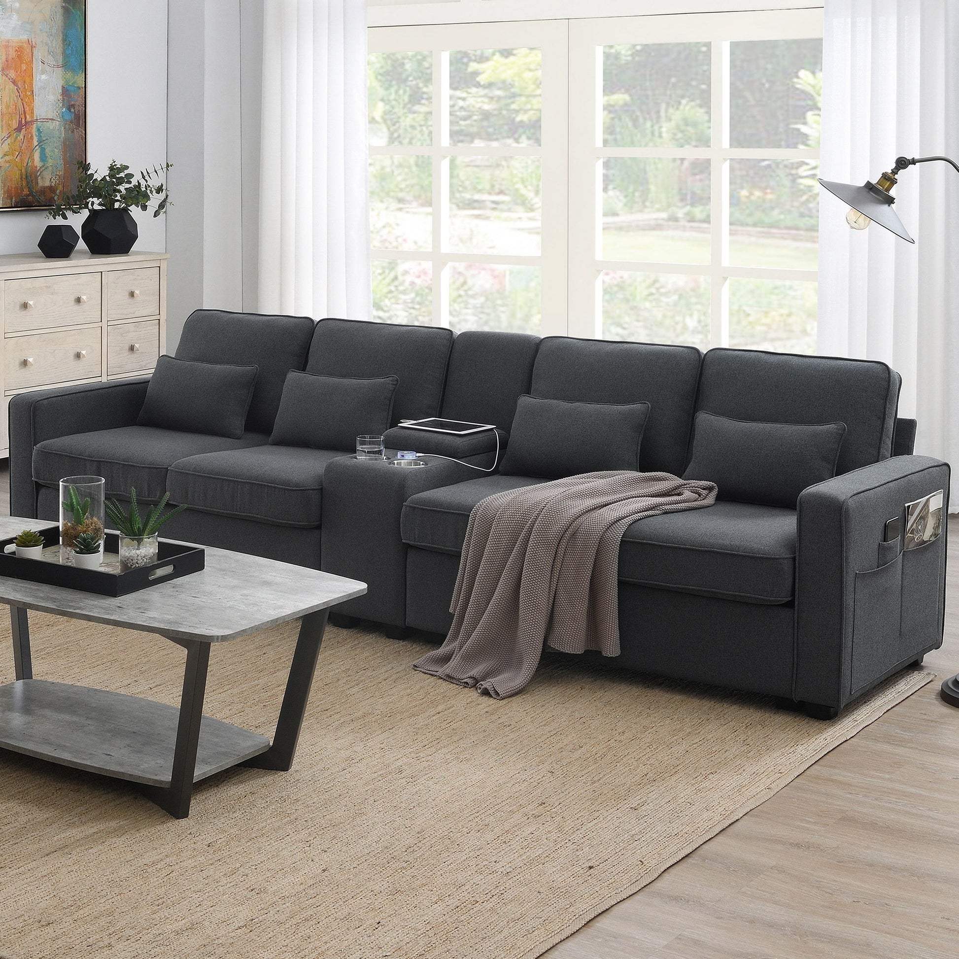 A modern living room with a dark gray upholstered 4-seat sofa featuring a console, 2 cupholders, 2 USB ports (wired/wireless charging), modern linen fabric, 4 pillows, a beige throw blanket, gray coffee table, floor lamp, dresser, and garden-view windows.