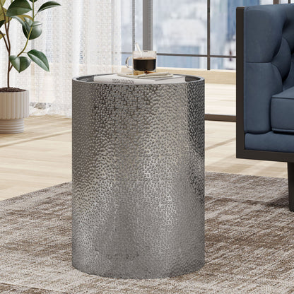 The Modern Round Accent Table With Hammered Surface, silver and stylish, holds a cup of coffee and a magazine. It stands on a light rug beside a dark blue sofa and plant in a bright living room with large windows.