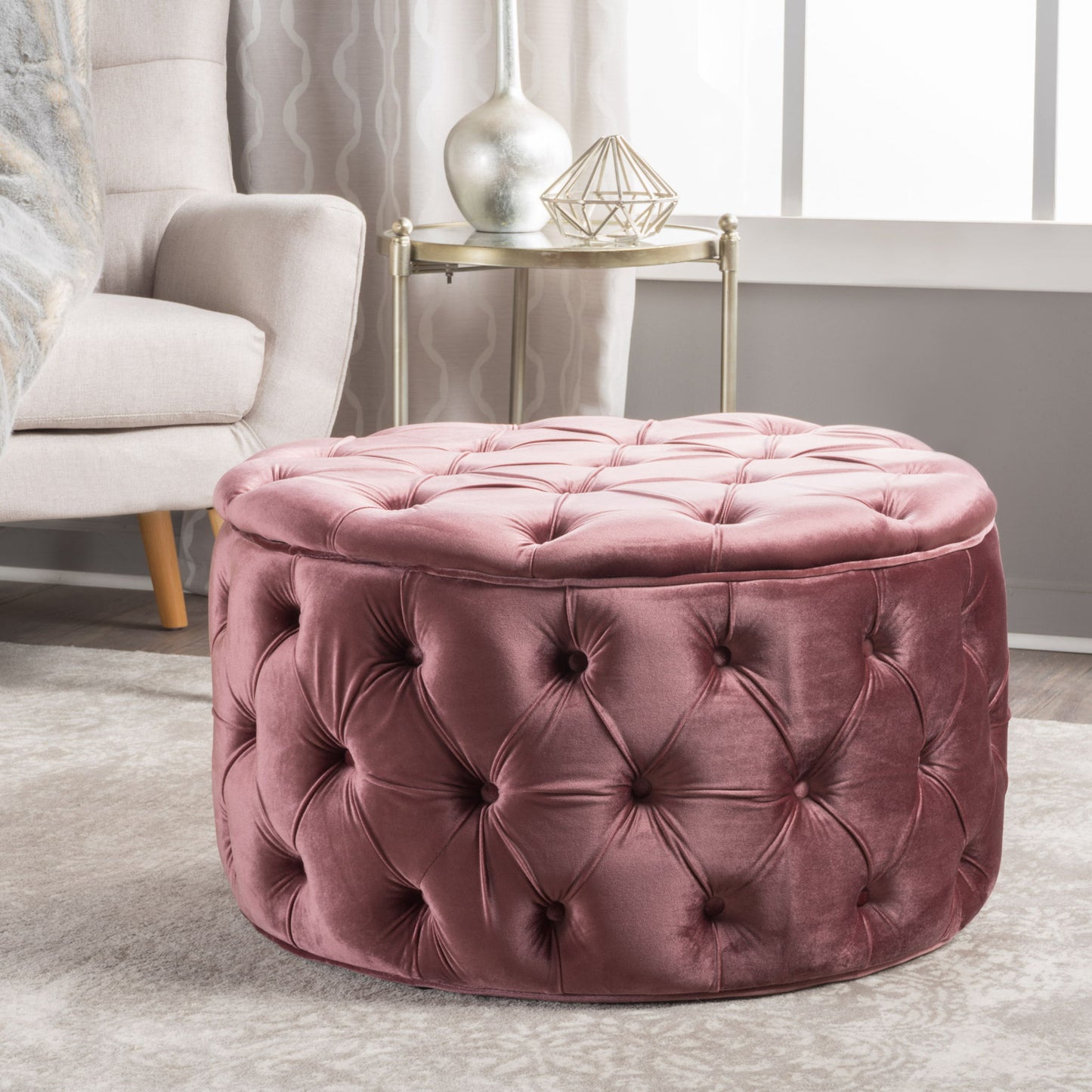 The Modern Glam Ottoman Velvet, a round tufted pink piece, sits on a light patterned rug beside a beige armchair and small side table with decor in a bright, modern living room.