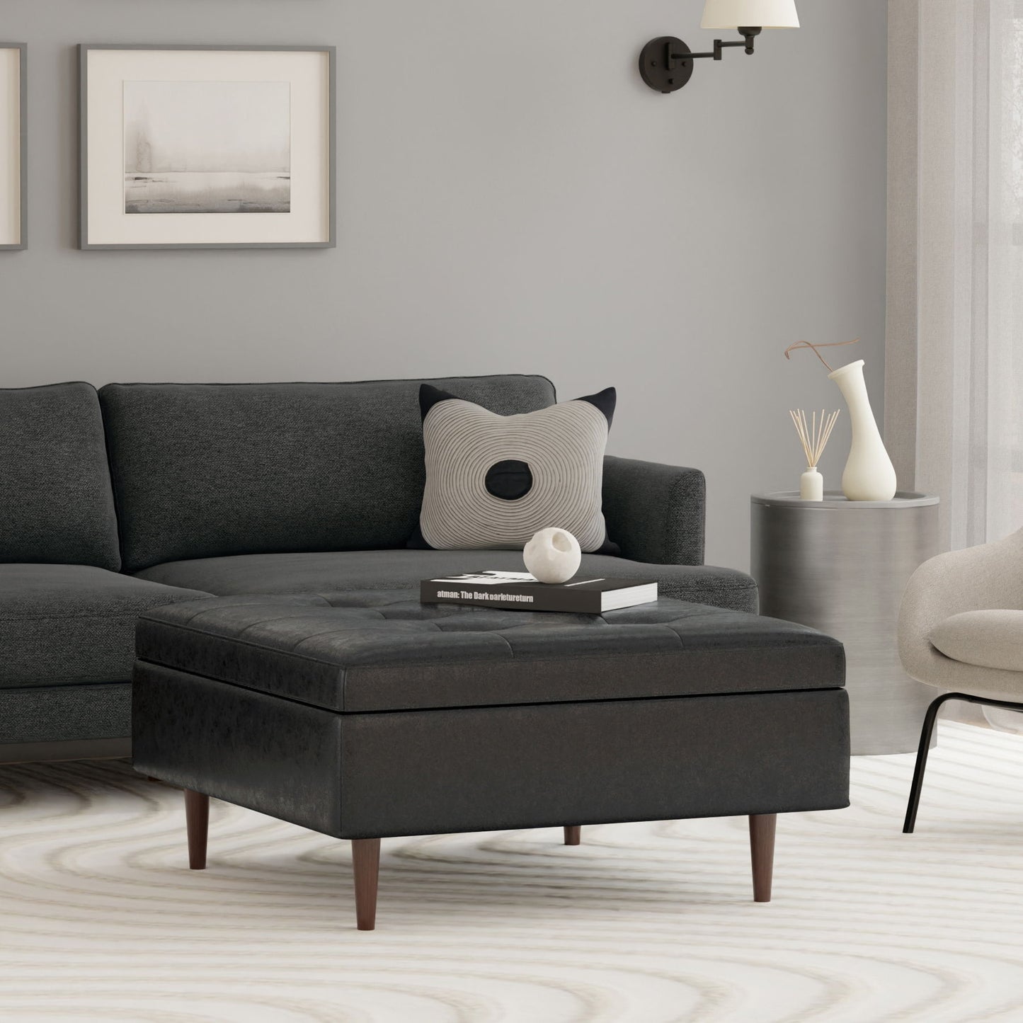A modern living room features a dark gray sectional, a patterned pillow, abstract wall art, a neutral rug, a round side table with decor, and the Shay Large Square Coffee Table Storage Ottoman in mid-century style with wooden legs.