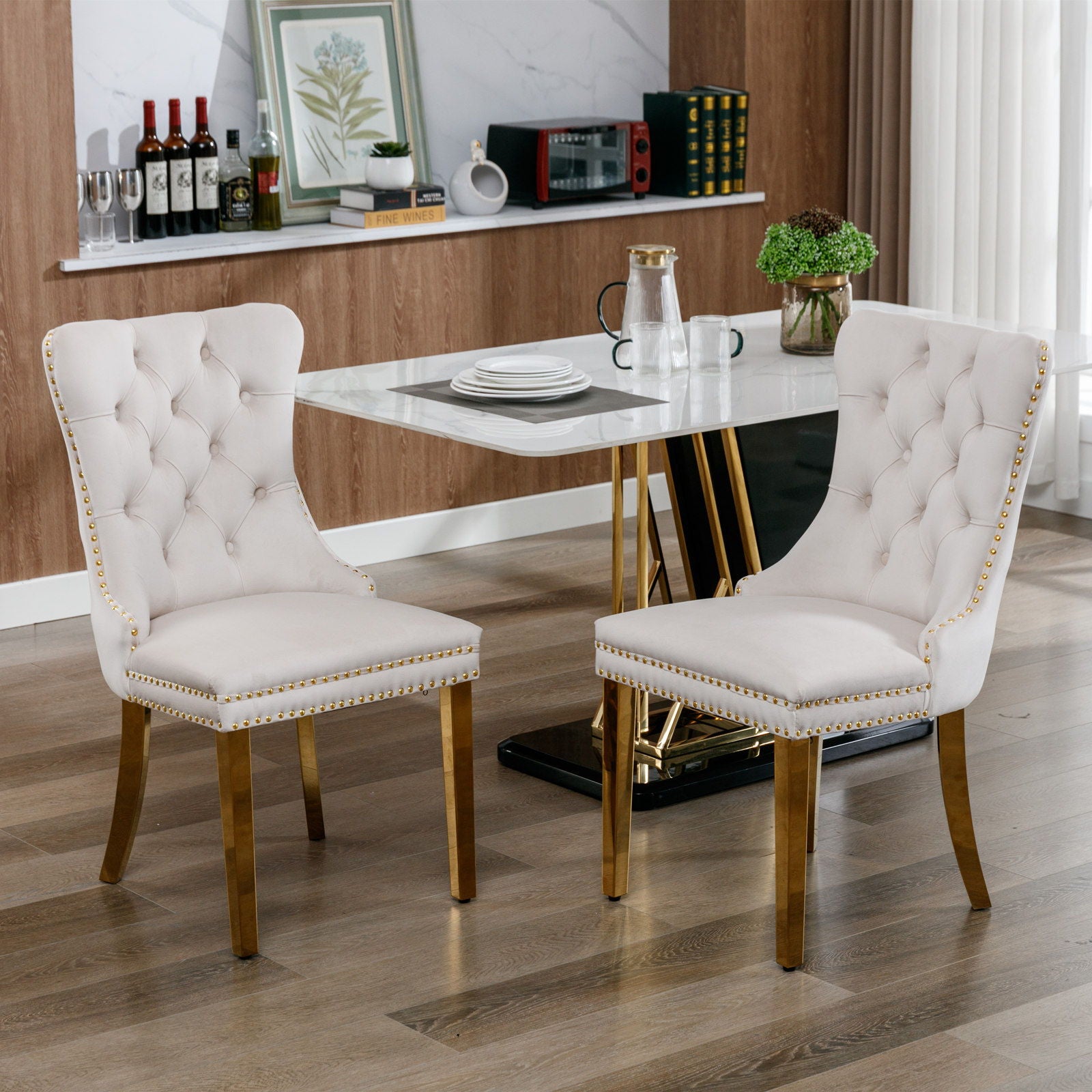 Two Nikki modern tufted velvet dining chairs with golden stainless steel legs and nailhead trim sit on a wooden floor in a contemporary dining room, paired with a marble-top table set for a meal.