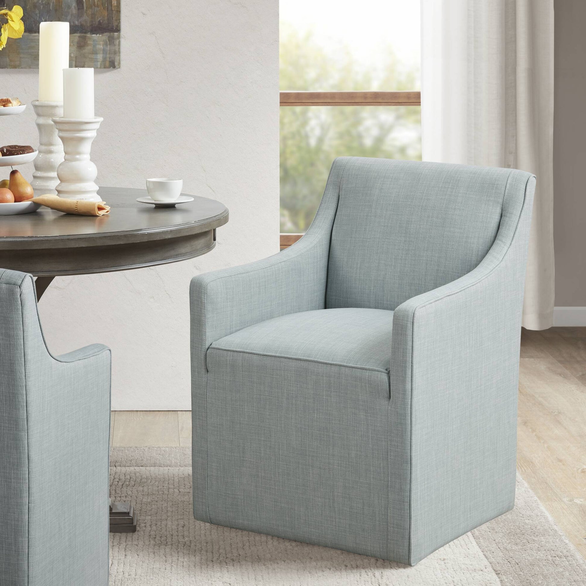 A Traditional Skirted Dining Arm Chair with light blue upholstery and a modern design sits by a round dining table set with white candles, pastries, and a cup in a bright room featuring large windows and neutral decor.