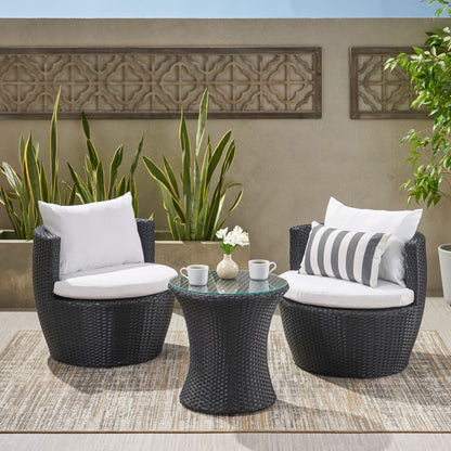 The Kono - Chat Set Outdoor Furniture features two black wicker chairs with white cushions on a rug beside a matching round table, topped with two mugs and a vase of flowers, set against green plants and a decorative wall.