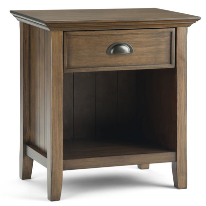 The Acadian Handcrafted Bedside Table is a wooden nightstand with a single drawer featuring a metal handle and an open lower shelf, displayed at an angle against a white background.