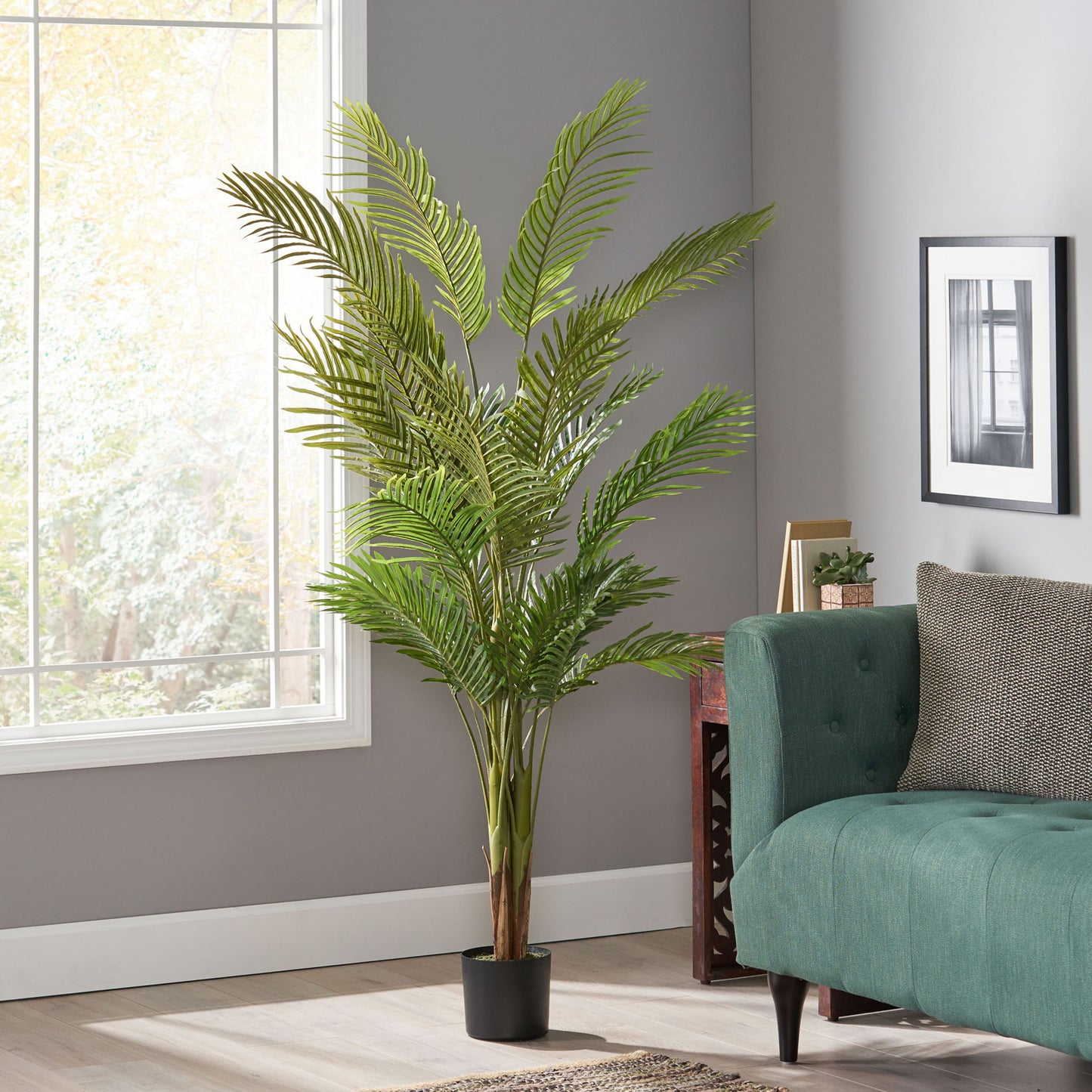 An Artificial Palm Tree in a black pot stands beside a green sofa in a modern living room with gray walls, a large window, and framed artwork.