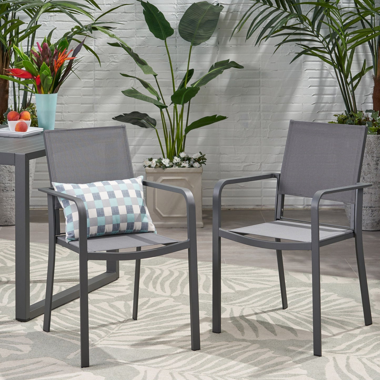 Two Waterfal Outdoor Aluminum Dining Chairs with mesh seats sit on a patterned rug. One chair has a checkered pillow. A small table with fruit and flowers is nearby, and green plants are set against a white brick wall in the background.