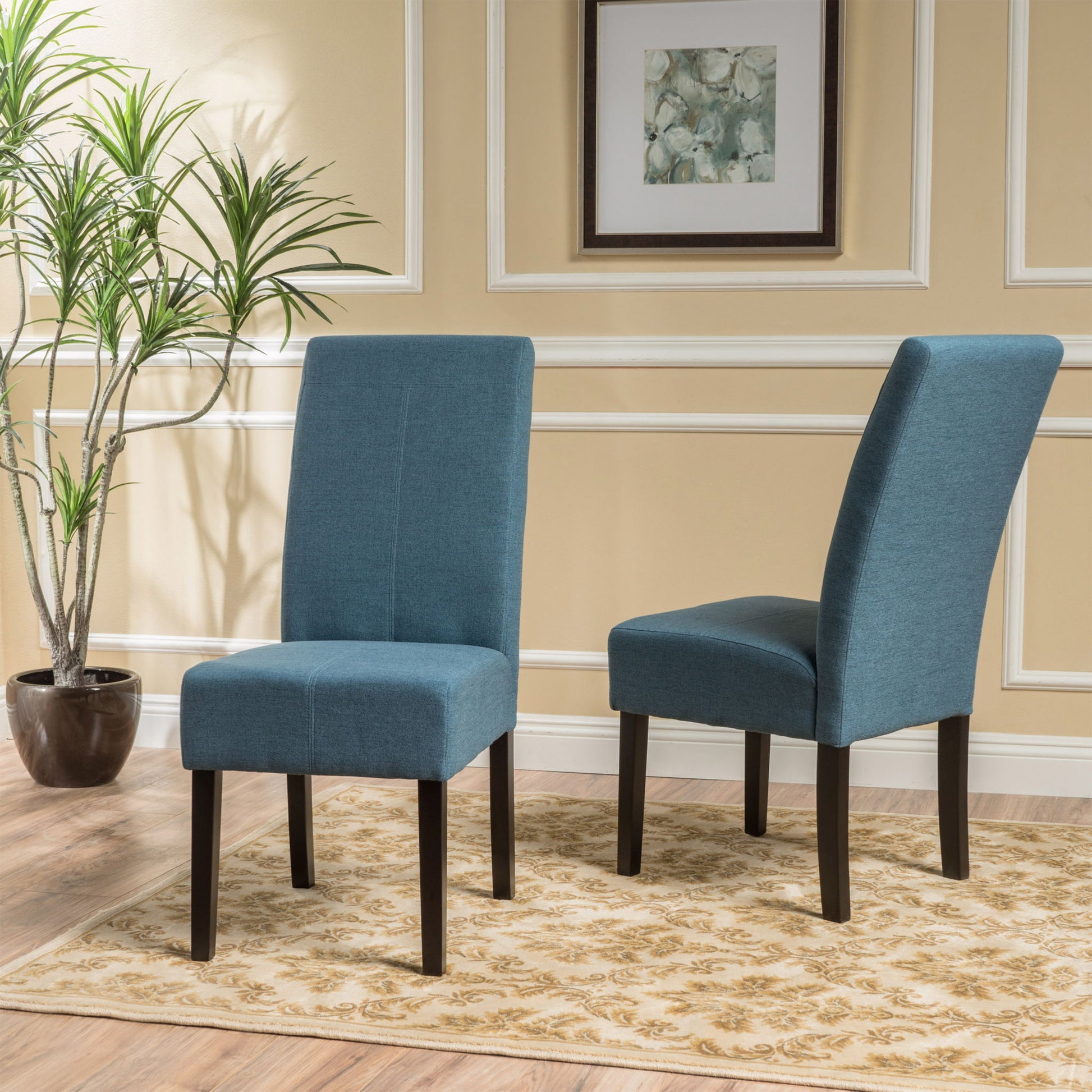 A set of two Pertica Dining Upholstered Armless Chairs with blue fabric and dark wooden legs sit on a light patterned rug in a beige room featuring white trim, framed art, and a potted plant.
