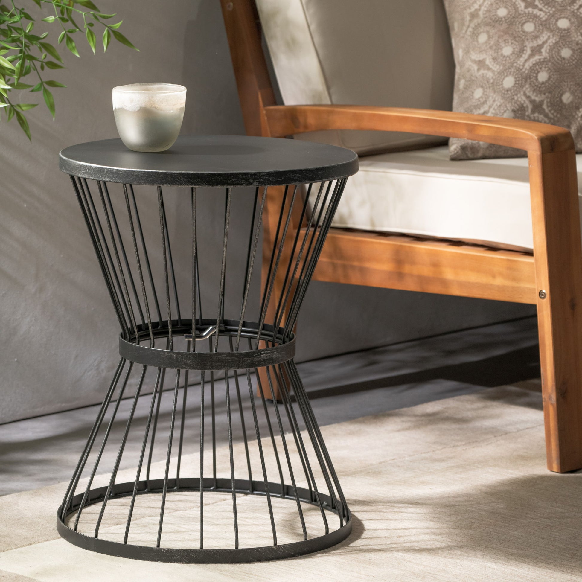 The Lassen Modern Outdoor Side Table with hourglass design, in sleek black, holds a candle beside a light-cushioned wooden armchair on a neutral rug in a cozy, sunlit room.