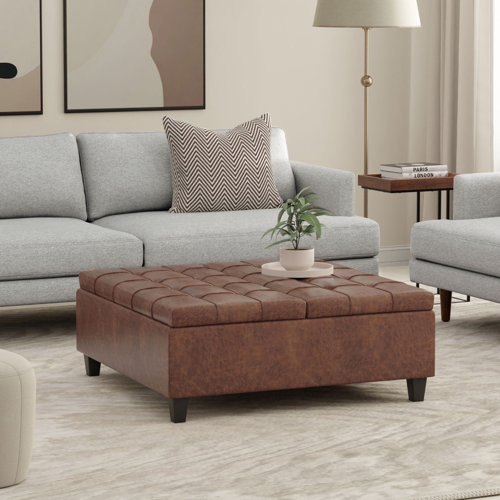 A modern living room with a light gray sofa, the Harrison Upholstered Coffee Table Storage Ottoman in brown, a potted plant atop it, abstract wall art, a side table with books, and a floor lamp—all in soothing neutral tones.