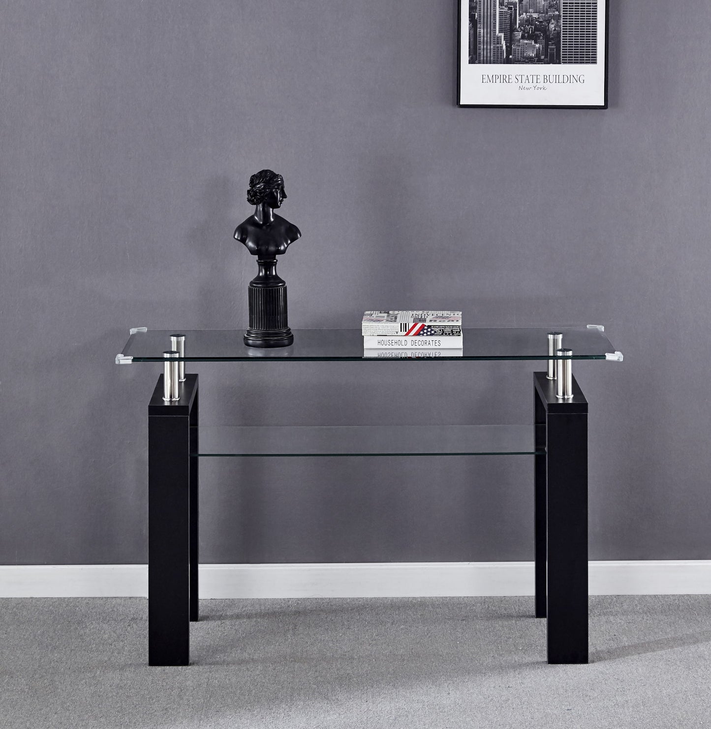The Clear Glass Top Side Table, Modern Design For Home, with black legs stands against a gray wall. On it sit a small black bust sculpture and two stacked books. A framed Empire State Building photo is mounted above.