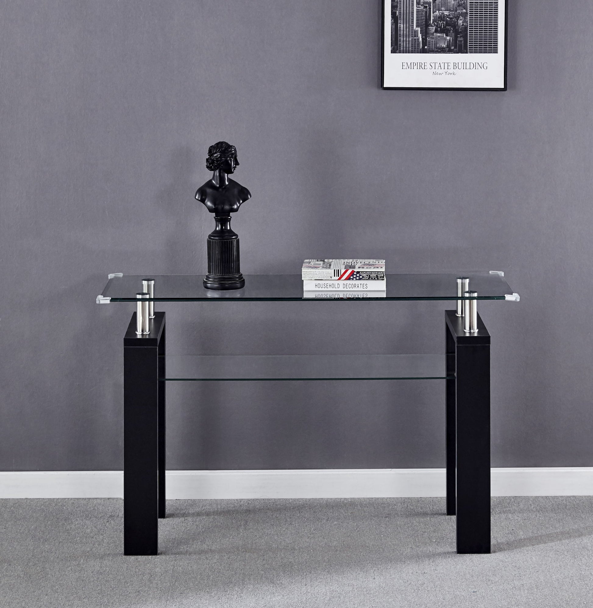 The Clear Glass Top Side Table, Modern Design For Home, with black legs stands against a gray wall. On it sit a small black bust sculpture and two stacked books. A framed Empire State Building photo is mounted above.