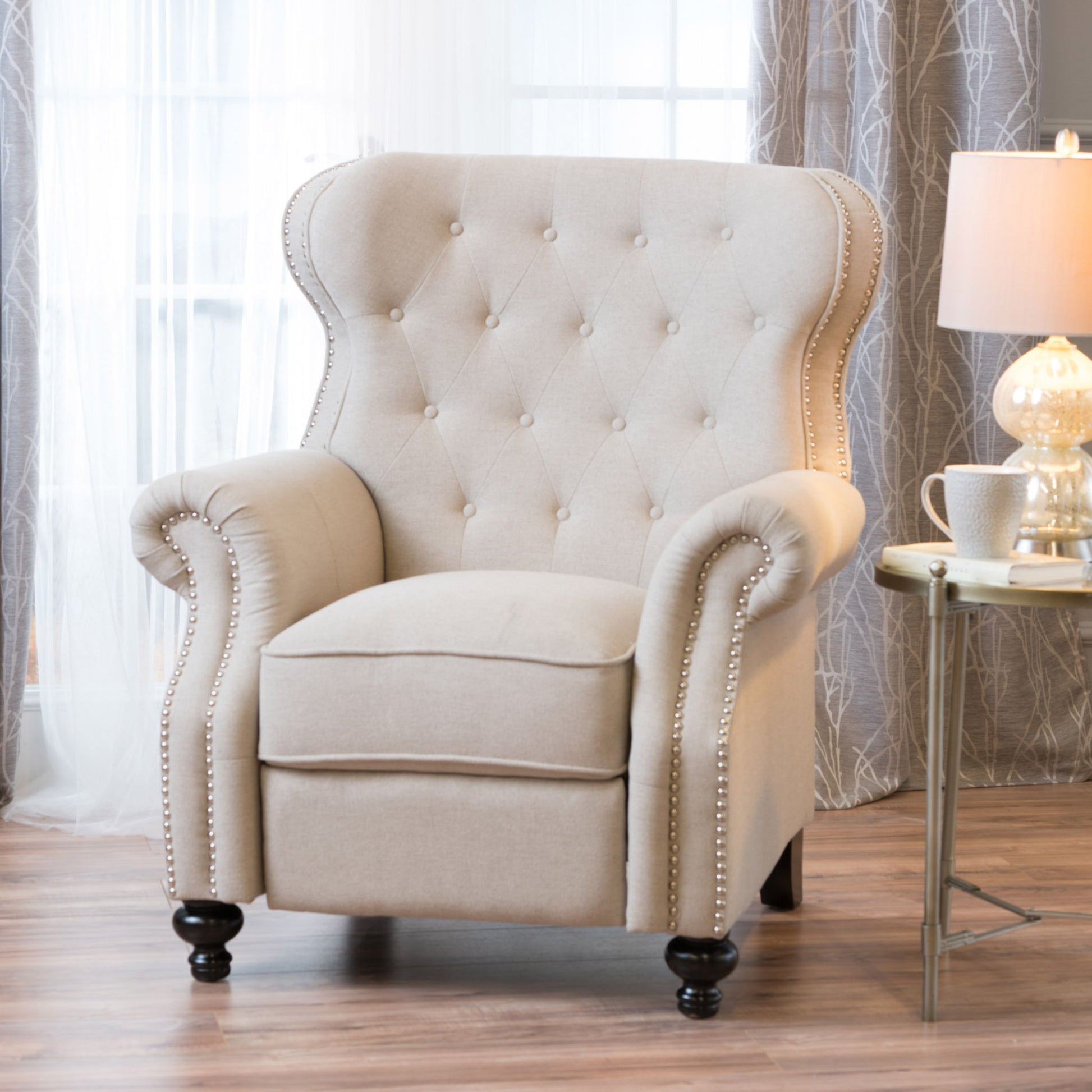 A Contemporary Style Push Back Recliner with button tufted stitching and nailhead trim sits on a wooden floor by a round side table holding a lamp, teacup, and book, in front of sheer curtains and a window.