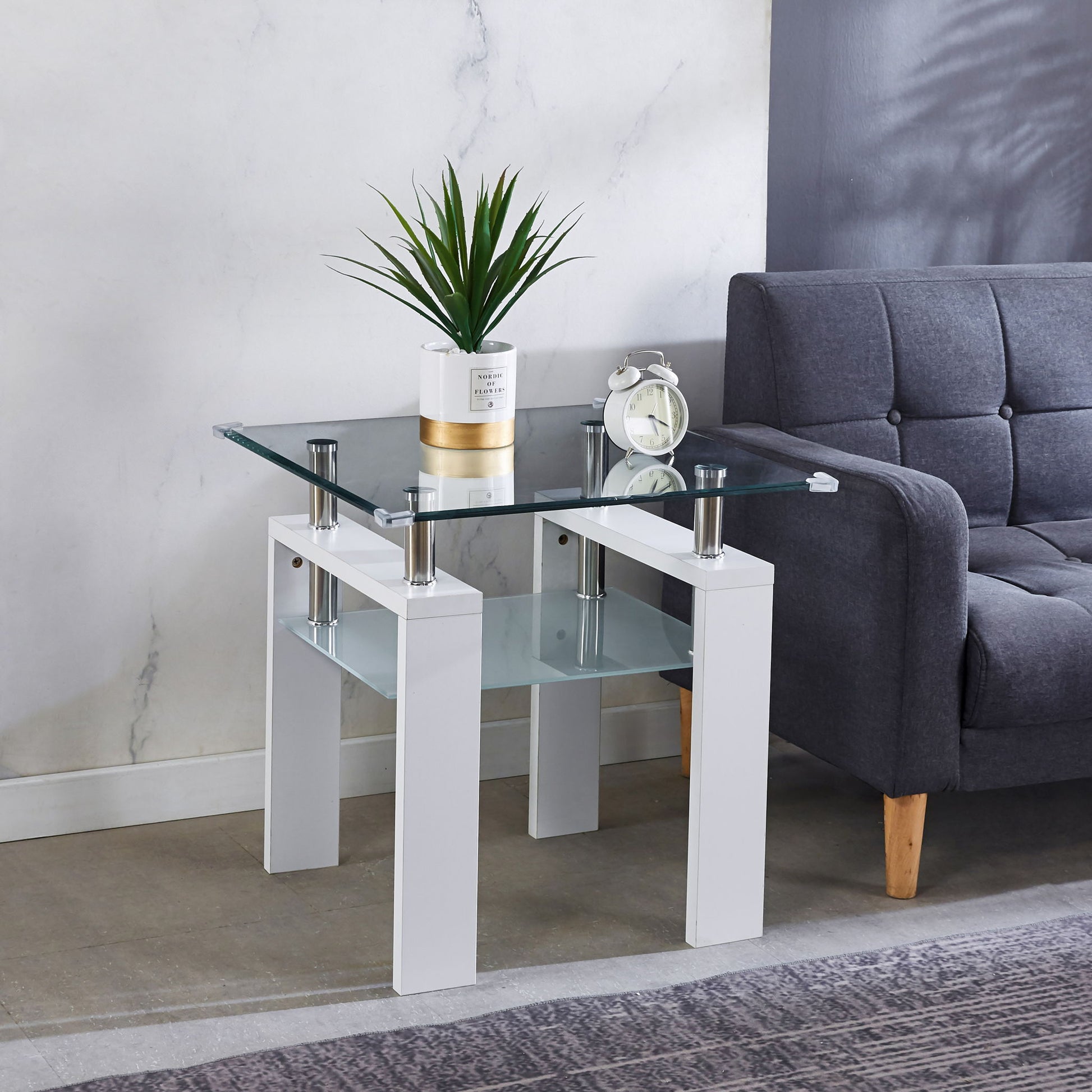 A modern living room features the Clear Glass Top Side Table, Modern Design For Home, with white legs holding a potted plant and a white alarm clock, placed beside a gray upholstered sofa with wooden legs.