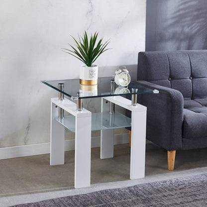 A modern living room features the Clear Glass Top Side Table, Modern Design For Home, with white legs holding a potted plant and a white alarm clock, placed beside a gray upholstered sofa with wooden legs.